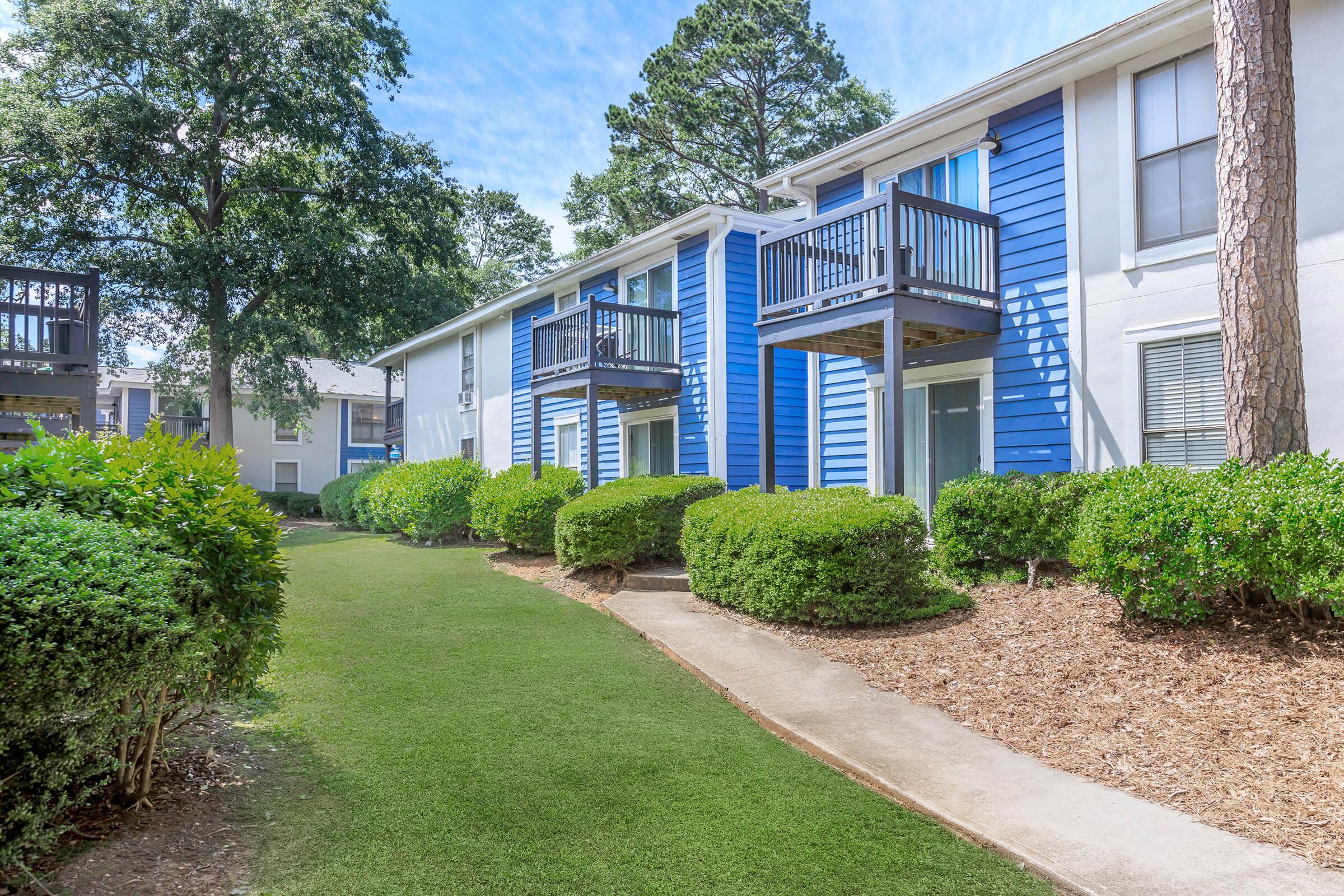 A well-maintained apartment complex with two-story buildings featuring blue siding. There are several balconies and neatly trimmed hedges lining a grassy pathway. Trees provide shade, and the sky is clear, suggesting a bright, sunny day.