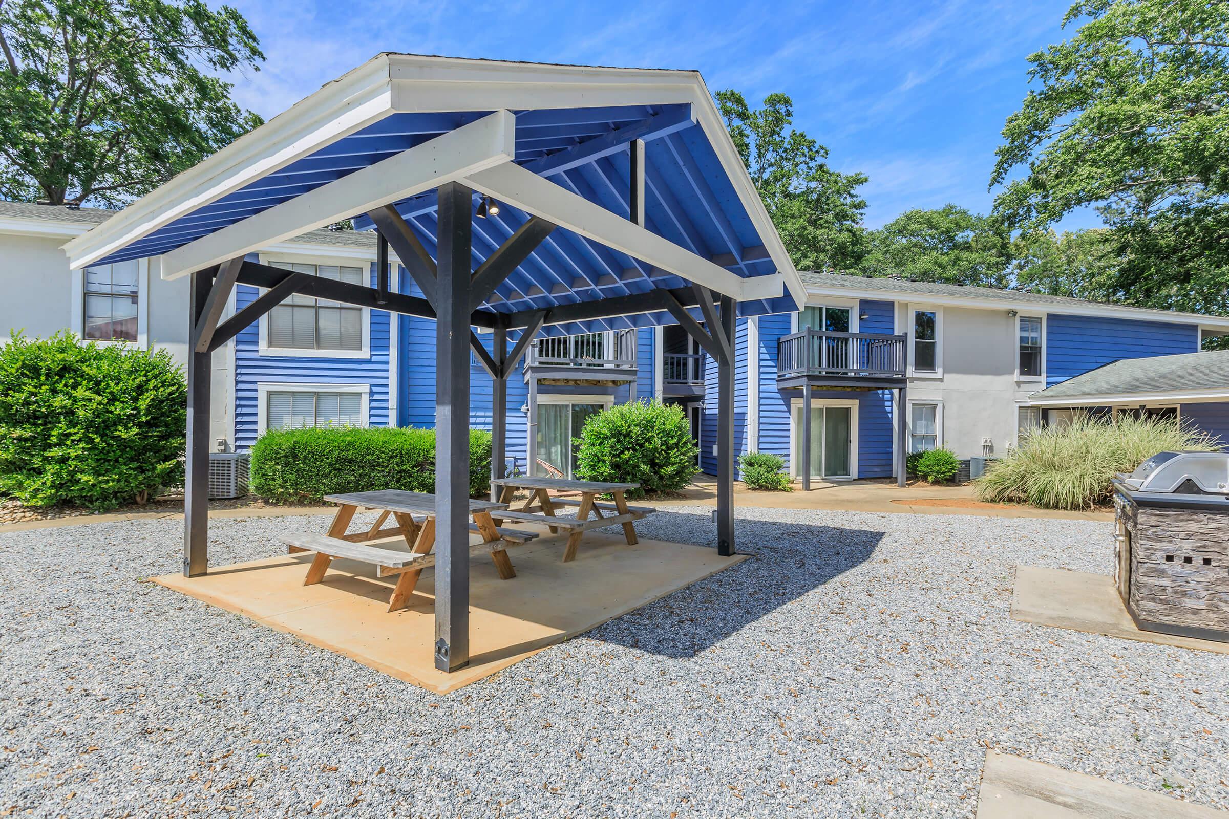 A shaded picnic area with a blue roof and wooden picnic tables sits on a gravel surface. Surrounding the area are buildings painted in blue and white, with balconies and greenery. The scene is bright and inviting, with clear blue skies and trees in the background.