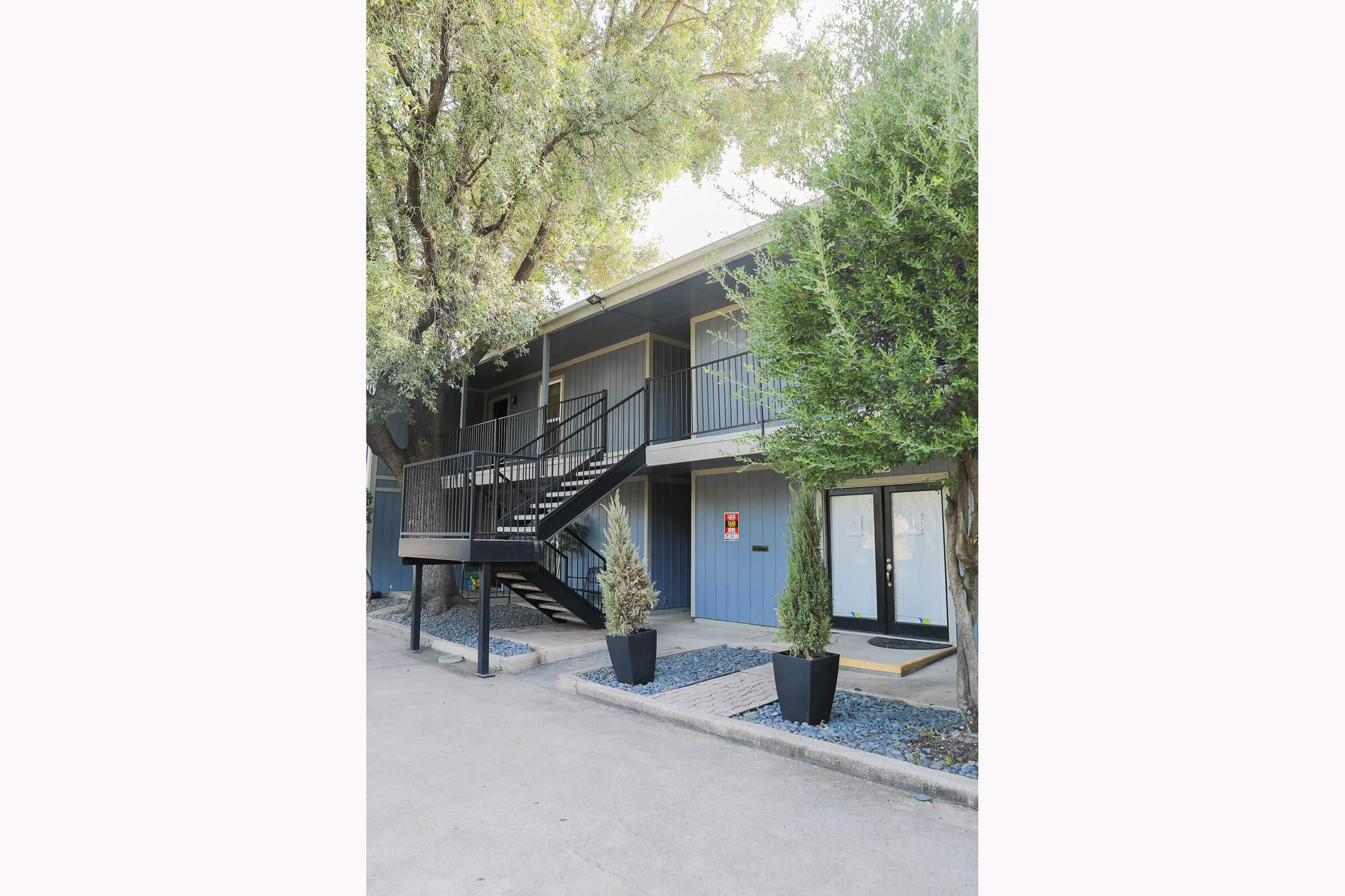 A two-story building with a grey exterior and a black metal staircase. The ground level features a glass door with an entry mat, flanked by two neatly trimmed shrubs. A pathway of decorative stones leads up to the entrance, surrounded by trees providing shade.