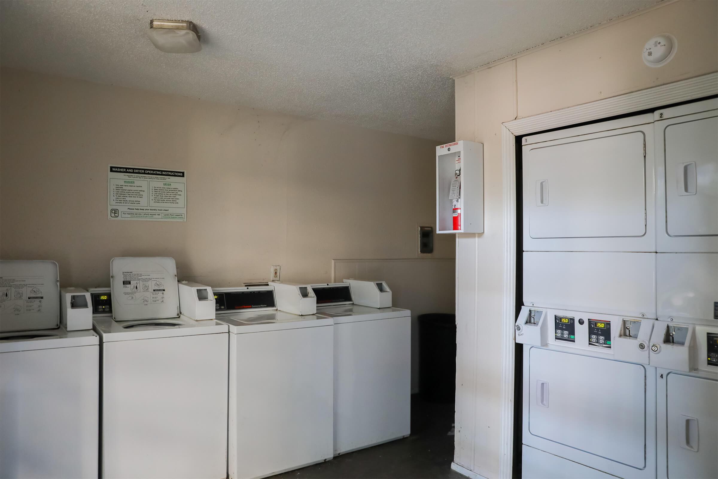 A laundry room featuring several white washing machines and a dryer. The walls are painted a light color, and there's a safety instructions sign on the wall. A vending machine is visible on the right side, and a black trash bin is located nearby. The room appears clean and organized.