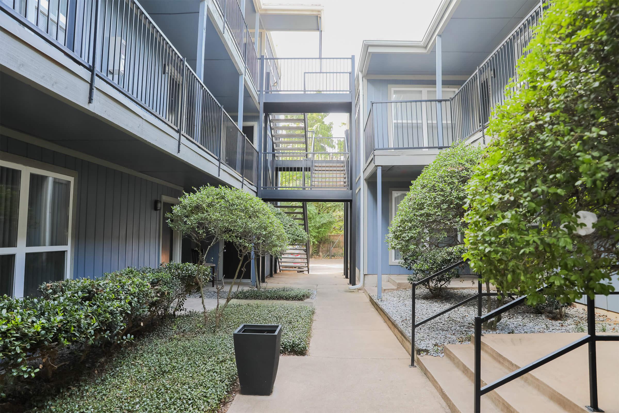 Courtyard view of an apartment complex featuring blue walls, green hedges, and trees. The scene includes a pathway leading to a staircase, various potted plants, and an outdoor seating area, creating a serene and inviting atmosphere.