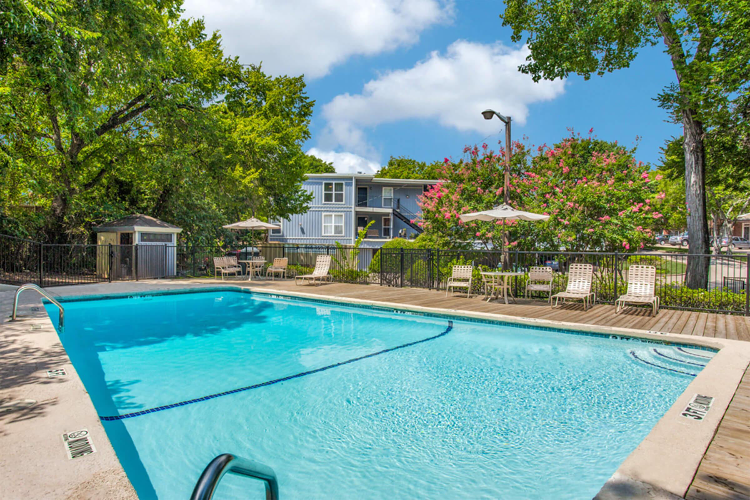 A clear swimming pool surrounded by wooden decking, lounge chairs, and umbrellas. Lush green trees and flowering plants are visible in the background under a blue sky with fluffy clouds. A small shed or structure is also present near the pool area.