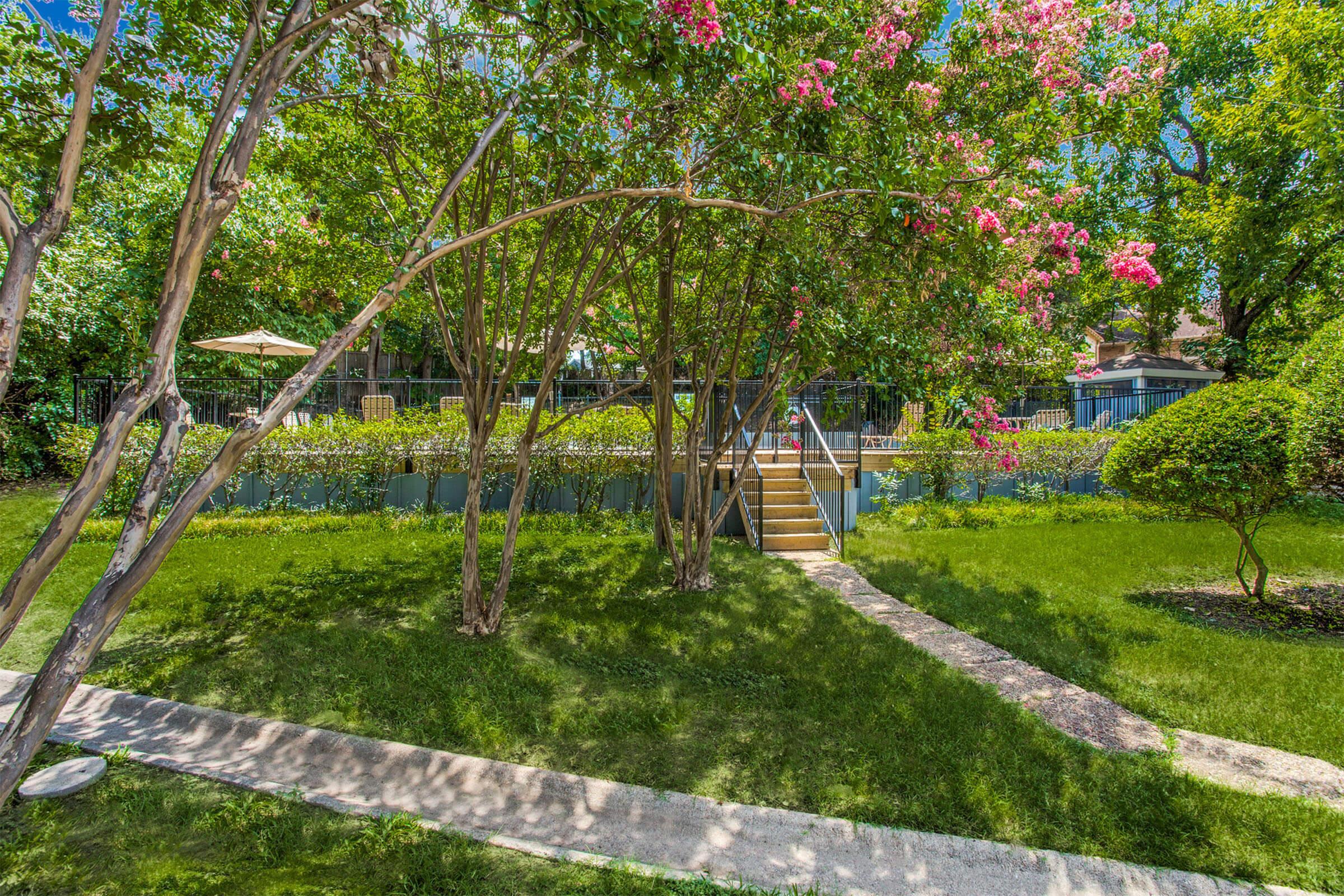 Lush garden scene featuring vibrant trees with pink flowers, a well-manicured lawn, and a stone path leading to wooden steps. In the background, a serene water feature and patio area with umbrellas are visible, surrounded by greenery under a clear blue sky.