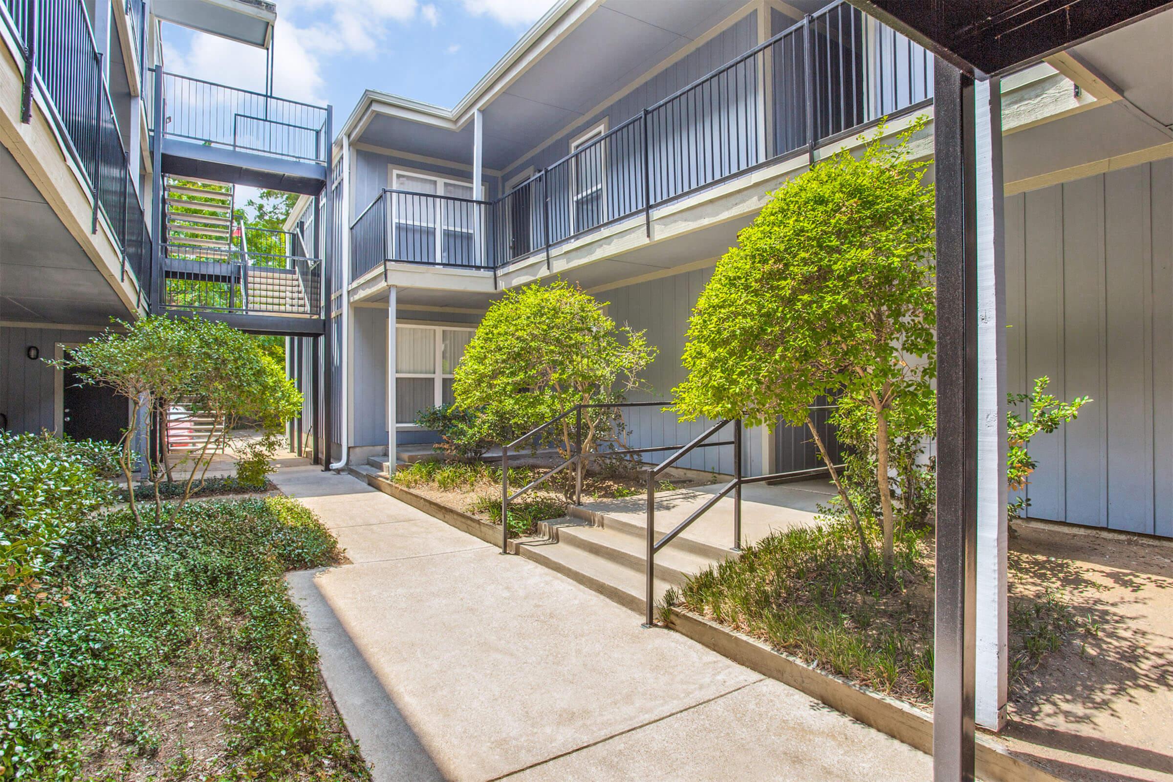 A view of an outdoor courtyard in an apartment complex featuring paved walkways, neatly trimmed shrubs, and greenery. The architecture includes multiple levels with balconies and stairs, surrounded by light-colored walls and railings. The scene is well-lit, indicating a sunny day.