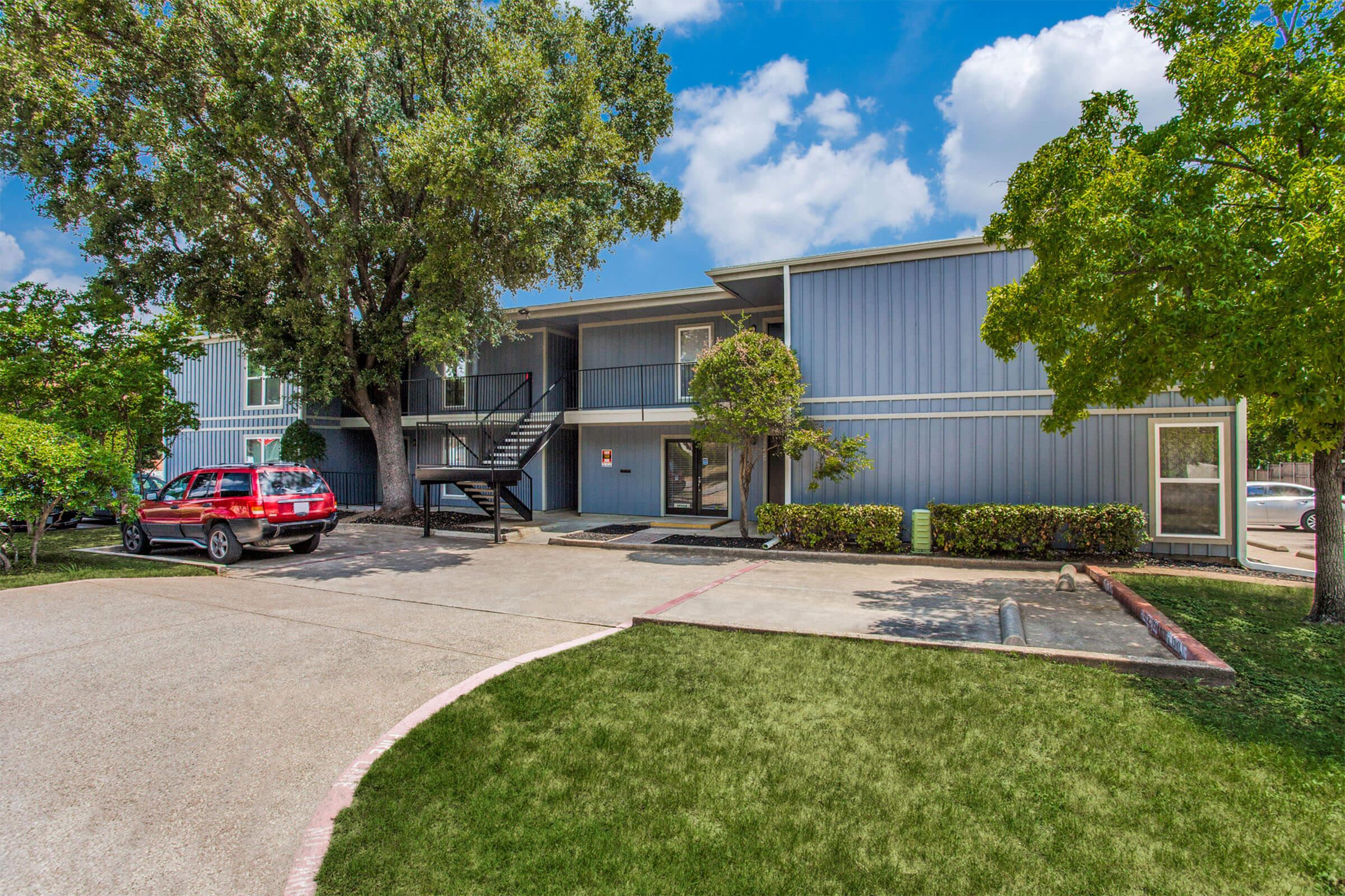A multi-unit apartment building with a blue exterior, featuring a staircase on the left side. The property has green trees and shrubs around, and a red car parked in the driveway. The sky is bright with scattered clouds, creating a sunny atmosphere. The lawn is well-kept and green.