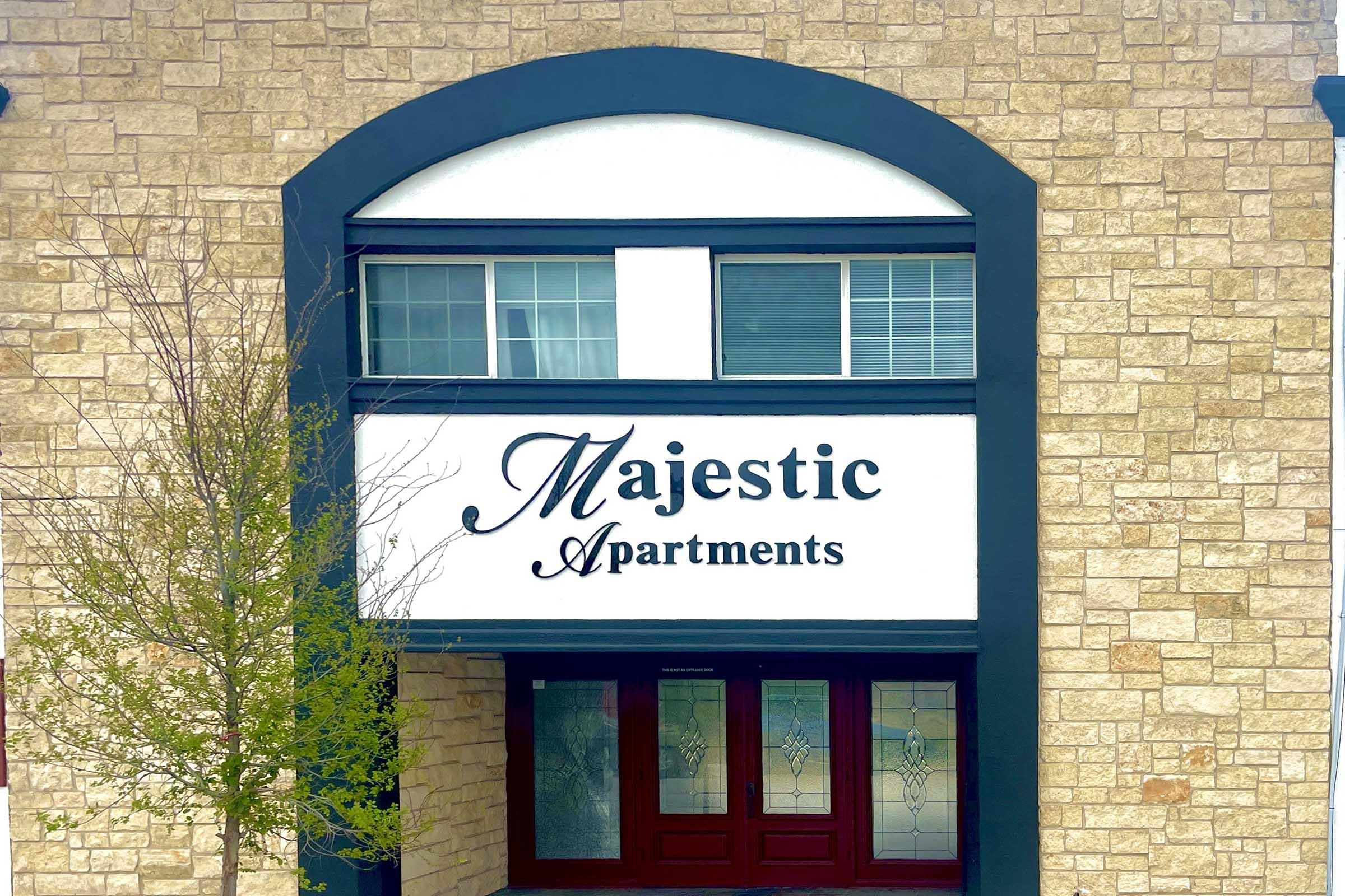 Sign for "Majestic Apartments" displayed on a stone building with large windows. The sign features elegant font against a white background, framed by a dark arch. A small tree is visible in front of the entrance, which has double wooden doors with decorative glass.