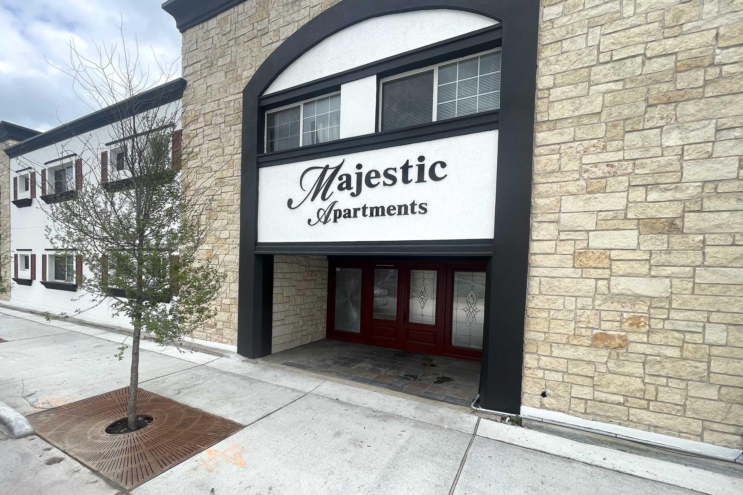 The image shows the entrance of a building with a sign that reads "Majestic Apartments." The façade features a mix of stone and painted surfaces, with large red doors and windows. A small tree is planted on the sidewalk in front of the entrance.