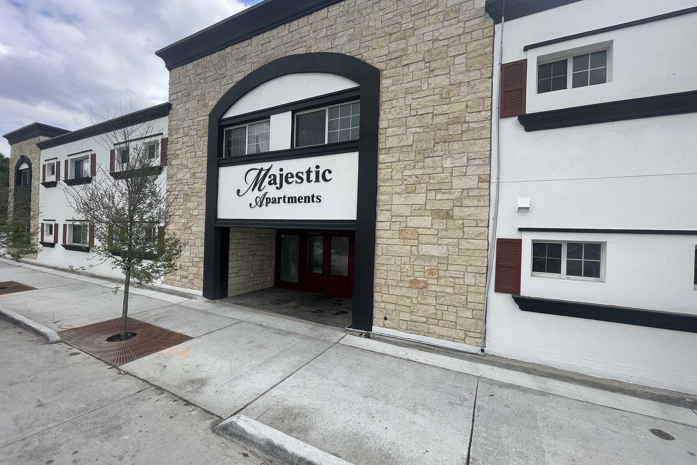 Exterior view of the Majestic Apartments building, featuring a stone facade and prominent entrance. The entrance has a large sign displaying "Majestic Apartments" and is flanked by red doors. The surrounding area includes sidewalks and small trees. Cloudy skies are visible above.