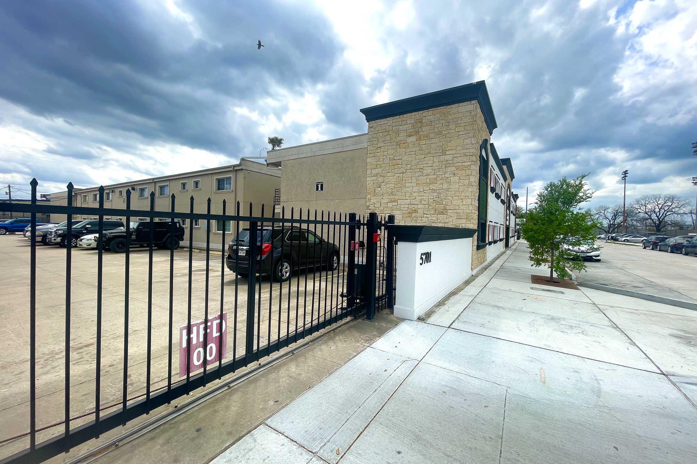 A view of a fenced area with a gate labeled "F1" and "S1," leading to a low-rise building. Parking spaces are visible, with several cars parked. The sky is cloudy, adding a moody atmosphere to the scene. A tree is planted in an area near the sidewalk.