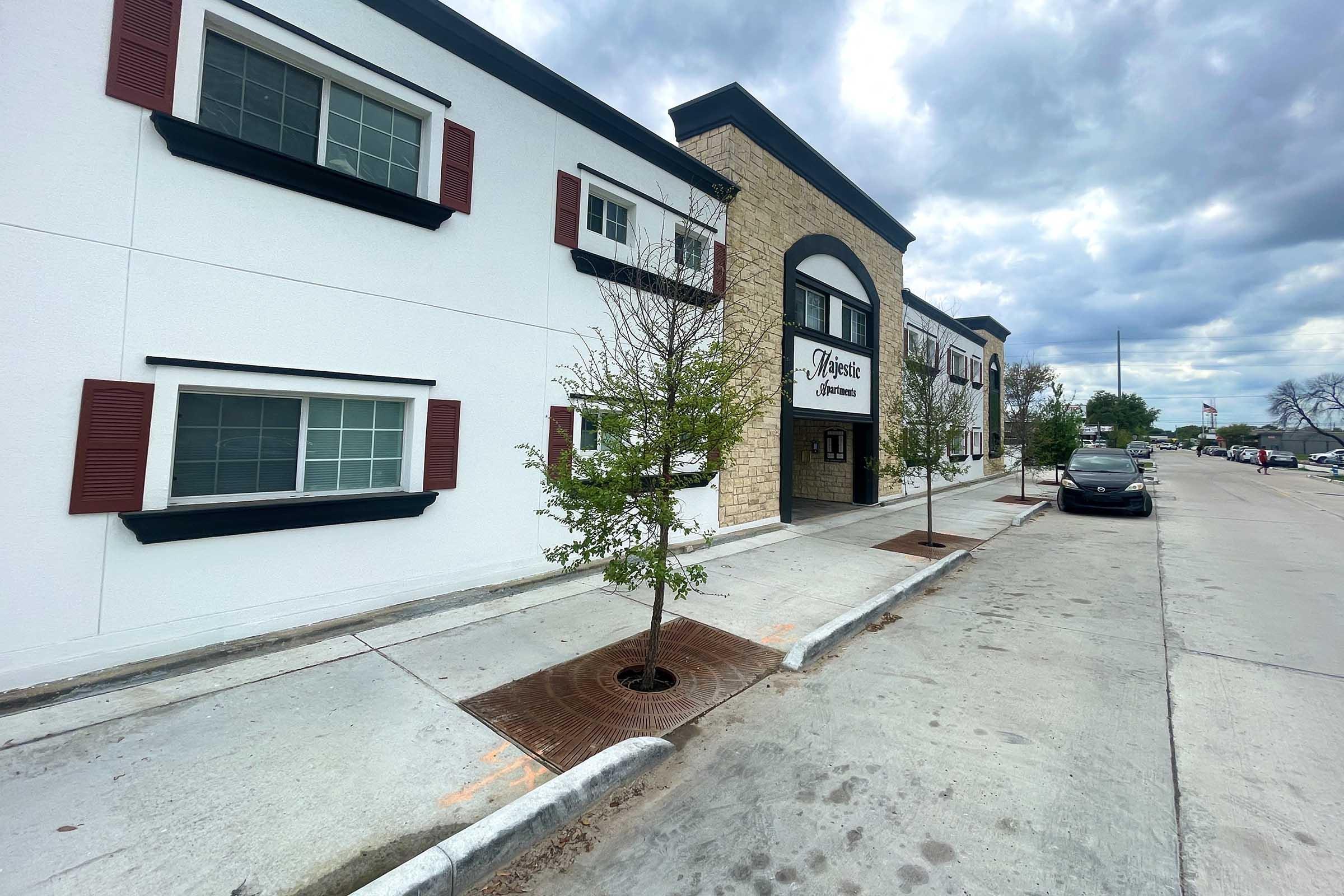 A modern two-story building with a white exterior and dark shutters, featuring a stone accent at the entrance. The entrance has a sign that reads "Majestic." Trees line the sidewalk, and a parked car is visible nearby. The sky is cloudy, hinting at overcast weather.