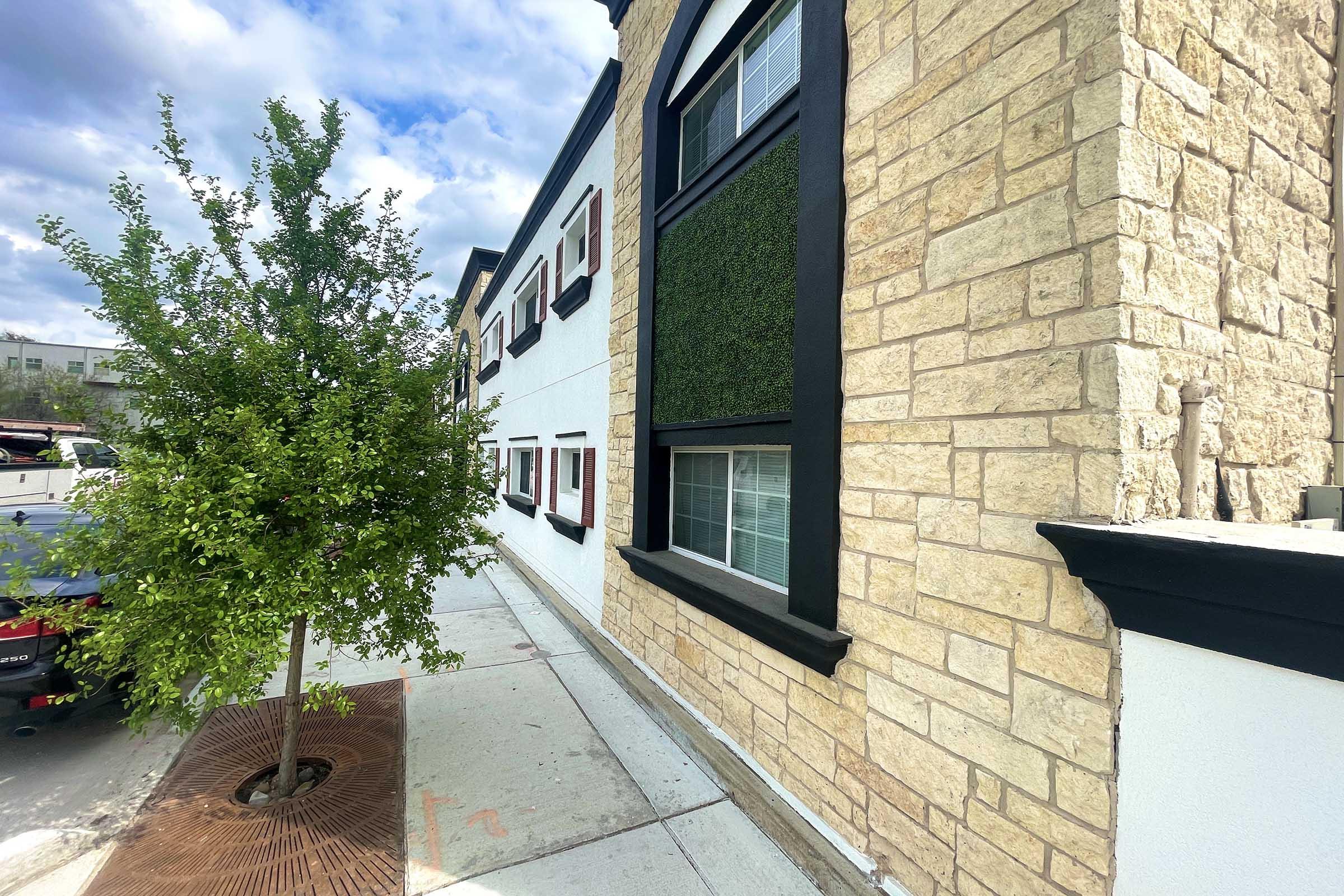 A view of a building's exterior featuring a stone wall, black trim, and windows with decorative elements. A small tree is planted near the sidewalk, and the scene is set under a partly cloudy sky.