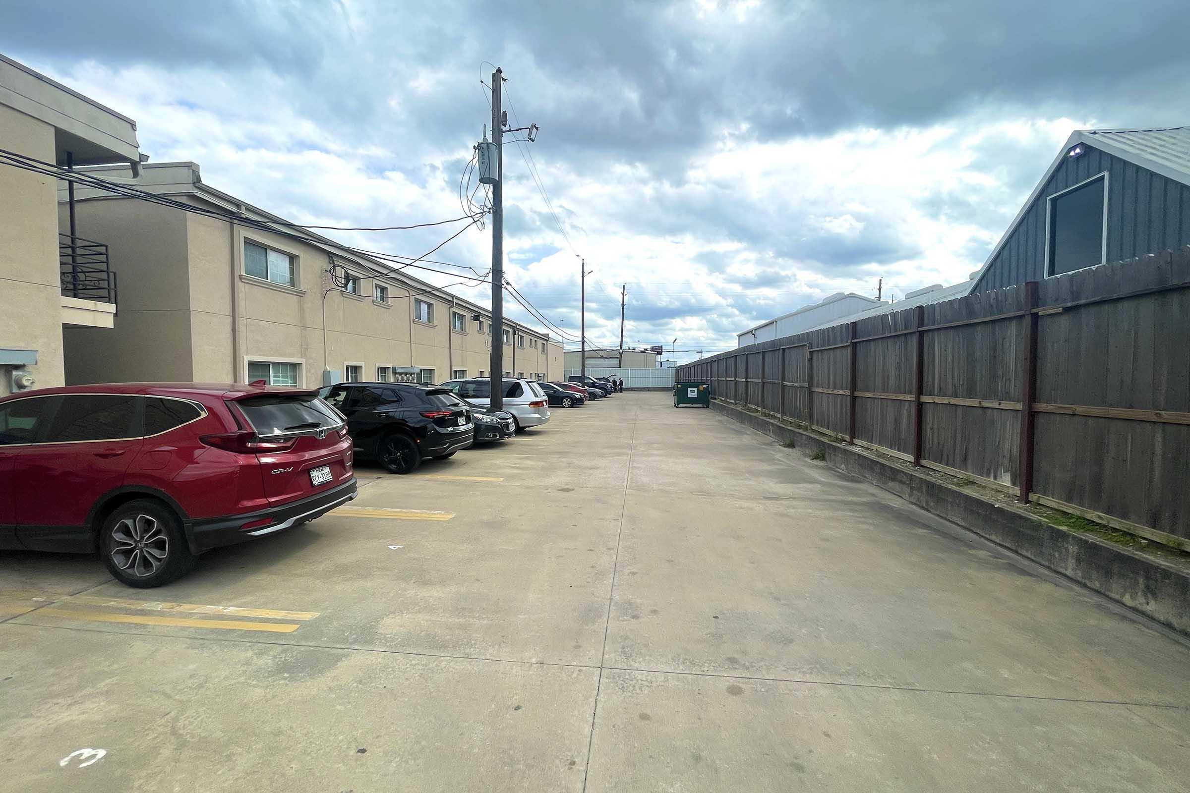 A view of a parking lot alongside a building, featuring several parked cars, including a red SUV and a darker sedan. In the background, a wooden fence and a dumpster are visible, with overcast skies above. The scene conveys a quiet, urban environment.