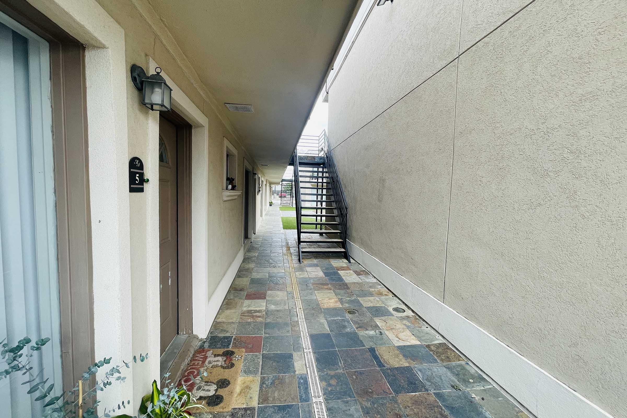 A narrow corridor between two buildings, featuring a tiled floor and light beige walls. On the left, there are two doors with numbers visible, and a staircase leading up on the right side. The area is well-lit, with greenery visible in the background.