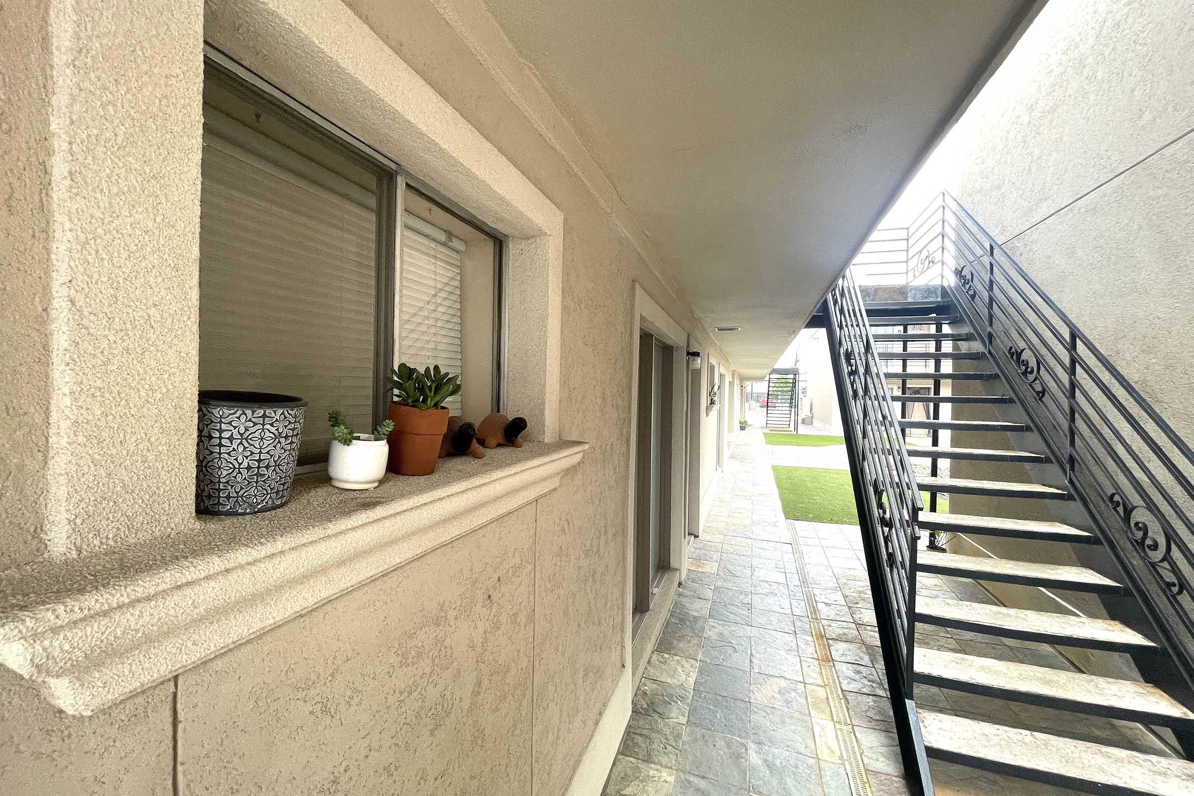 A view of a residential corridor featuring a row of windows with potted plants on the ledge. An exterior staircase with decorative railings is visible along the right side, leading up to an upper level. The floor is tiled, and there is a patch of grass at the end of the corridor, suggesting a well-maintained building.