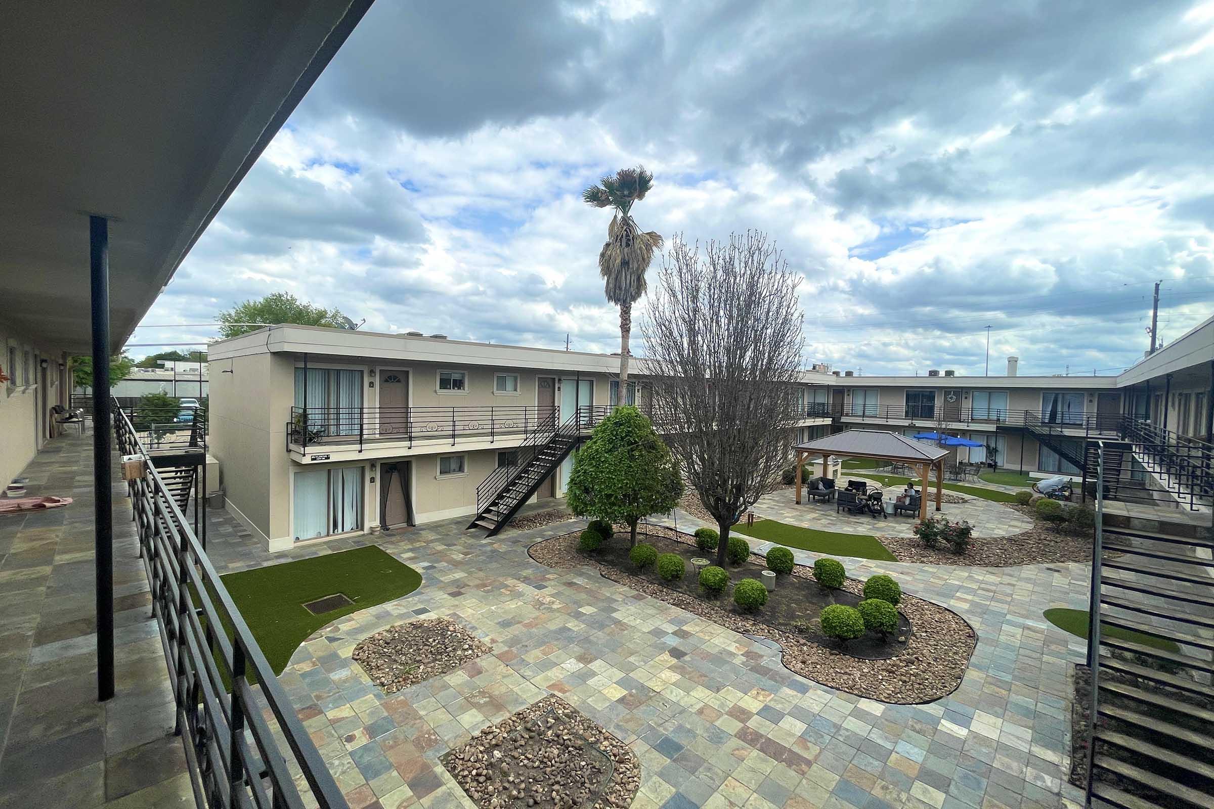 View of a courtyard in an apartment complex featuring two-story buildings. The area includes decorative stone pathways, neatly trimmed shrubs, a small palm tree, and a covered seating area. The sky is partly cloudy, adding to the tranquil atmosphere.