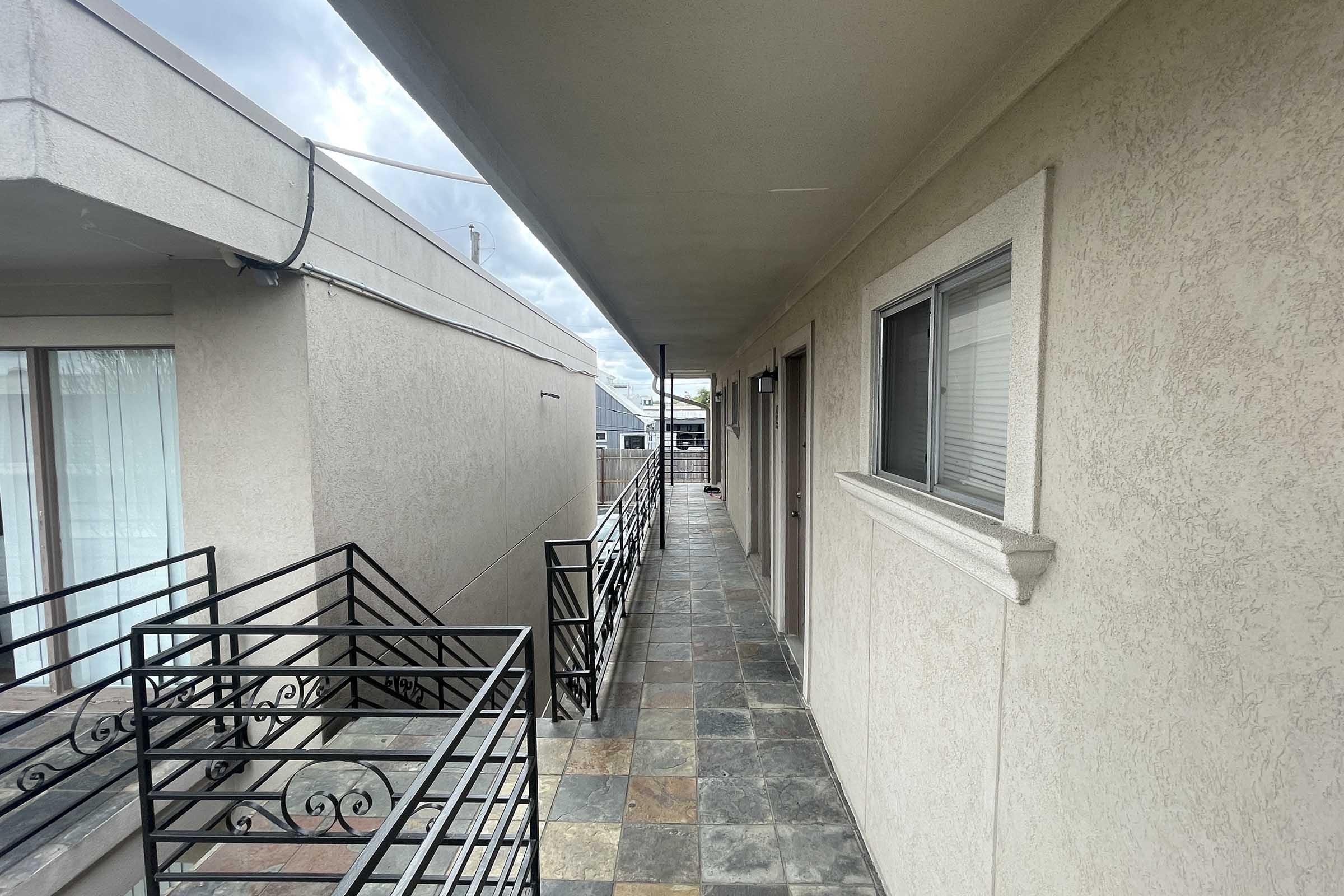 View of an exterior corridor with slate tiles and a railing. The corridor is flanked by doors on either side, with a cloudy sky visible in the background. The lighting is natural, illuminating the neutral-toned walls of the building.