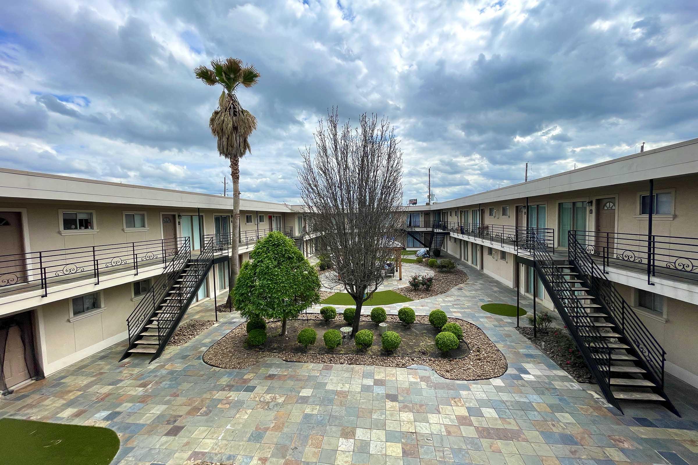 A view of a courtyard area in a low-rise motel or apartment complex. The scene features a mostly treeless area with a manicured lawn, decorative shrubs, and stone pathways. There are two levels of exterior walkways with staircases leading to individual units. The sky is cloudy, adding to the serene atmosphere.
