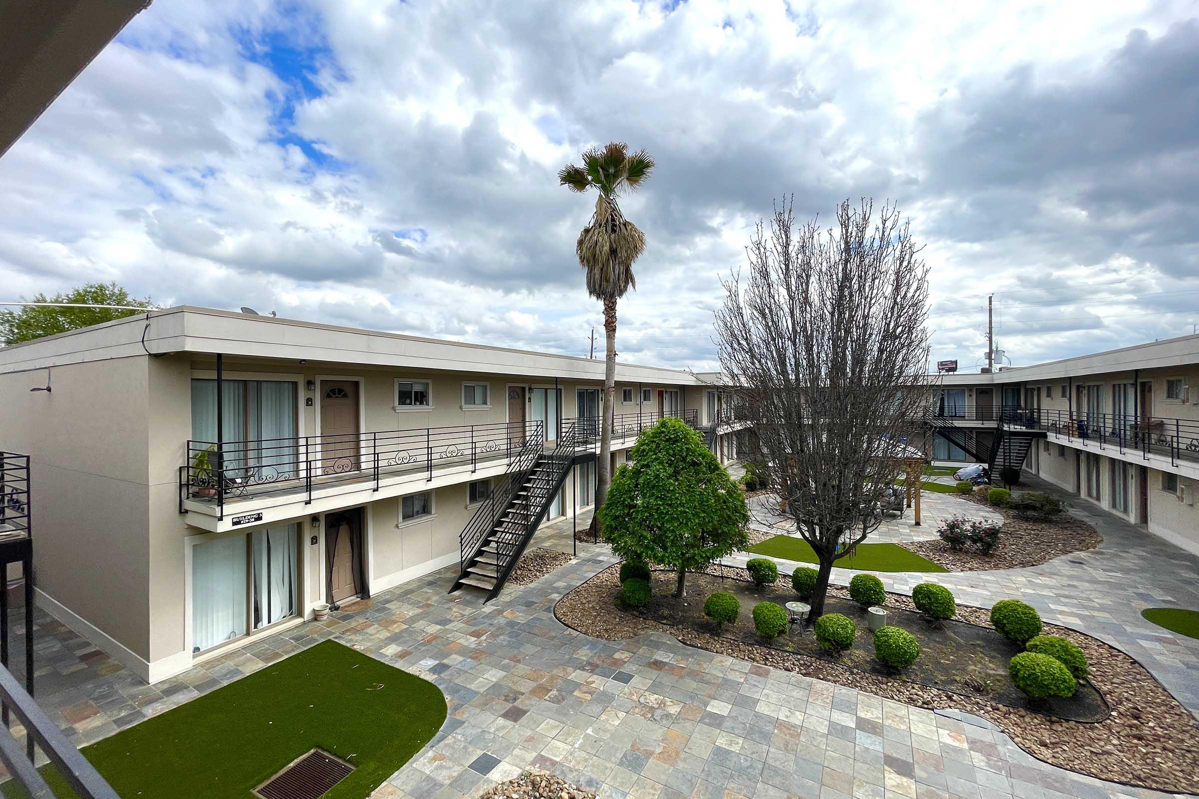 View of a motel courtyard featuring a landscaped area with shrubs and small trees, surrounded by two-story motel buildings. A palm tree stands in the center, and the cloudy sky is visible above. The scene showcases a modern, well-maintained outdoor space typical of motels.
