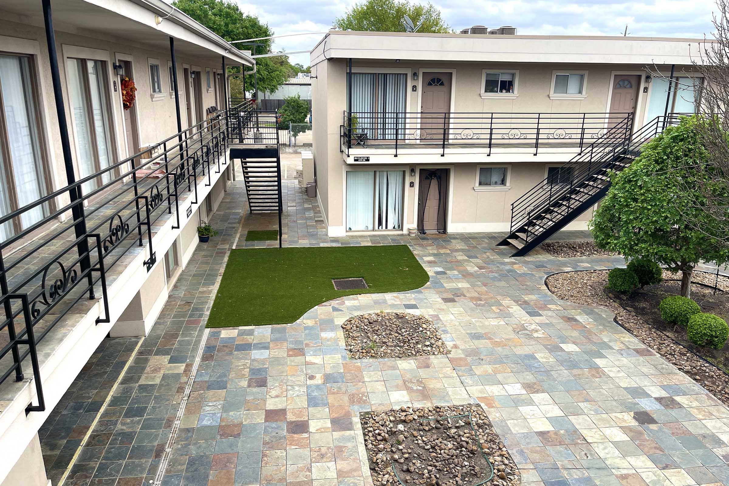 A view of a courtyard in an apartment complex featuring multiple levels of balconies. The courtyard has green grass, stone pathways, and landscaped areas with rocks and shrubs. There are stairs leading to the upper levels, and the buildings have a neutral color with windows and doors visible.