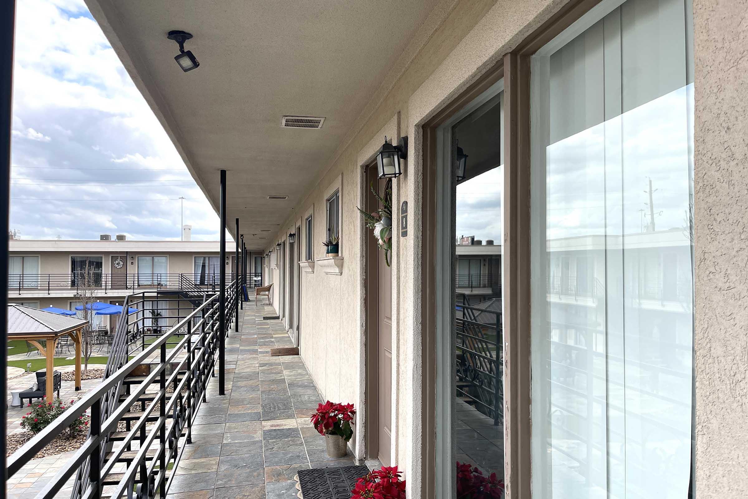 A view of a hotel corridor with doors on either side, featuring potted plants and holiday decorations. The floor is tiled, and the railing runs along with the walkway. In the background, another section of the building and a gazebo can be seen under a cloudy sky.