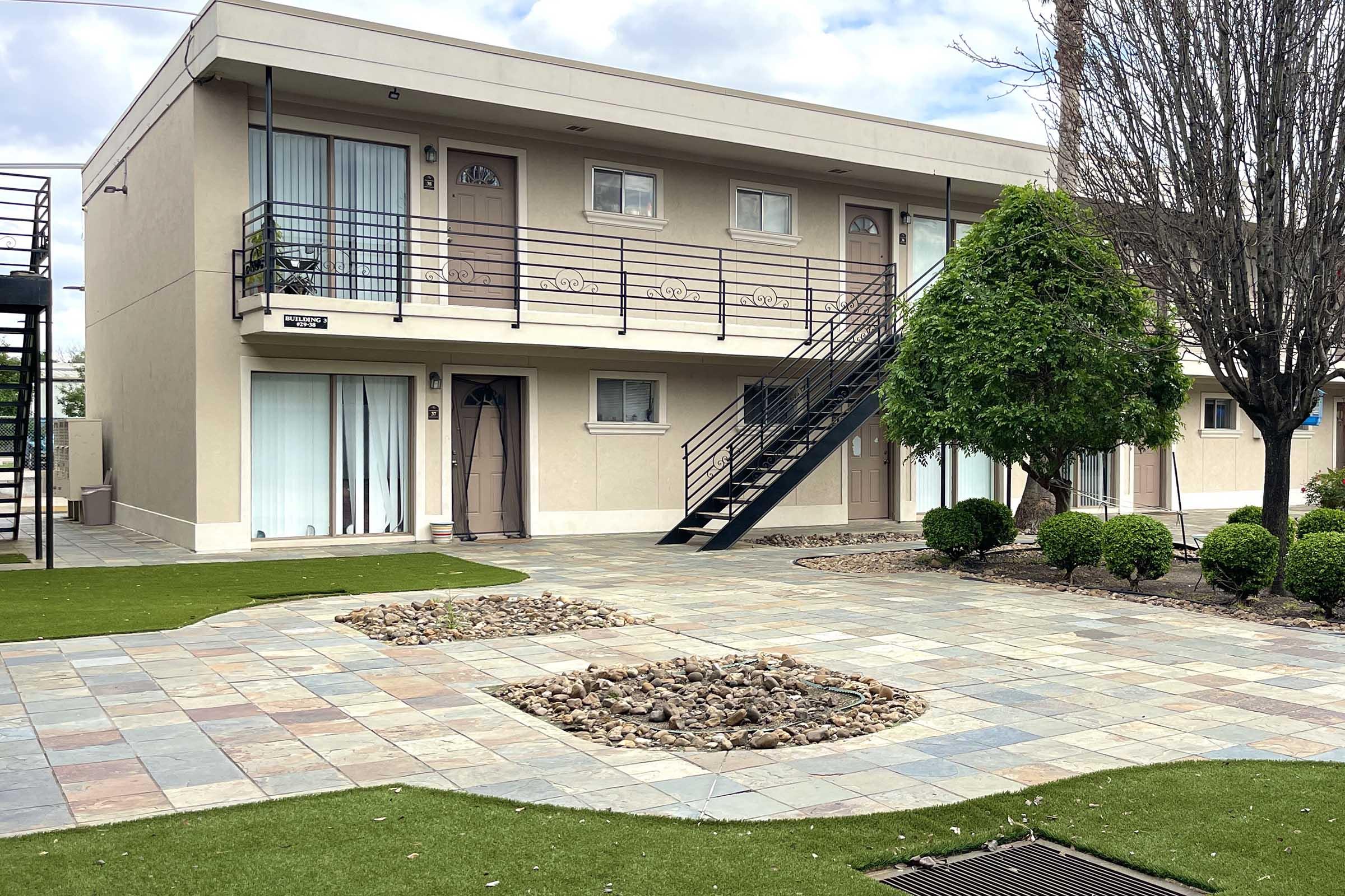 A two-story building featuring multiple doors and balconies, surrounded by manicured greenery and a landscaped pathway with small stones. The building has a modern design with large windows and railing stairs leading to the second floor.