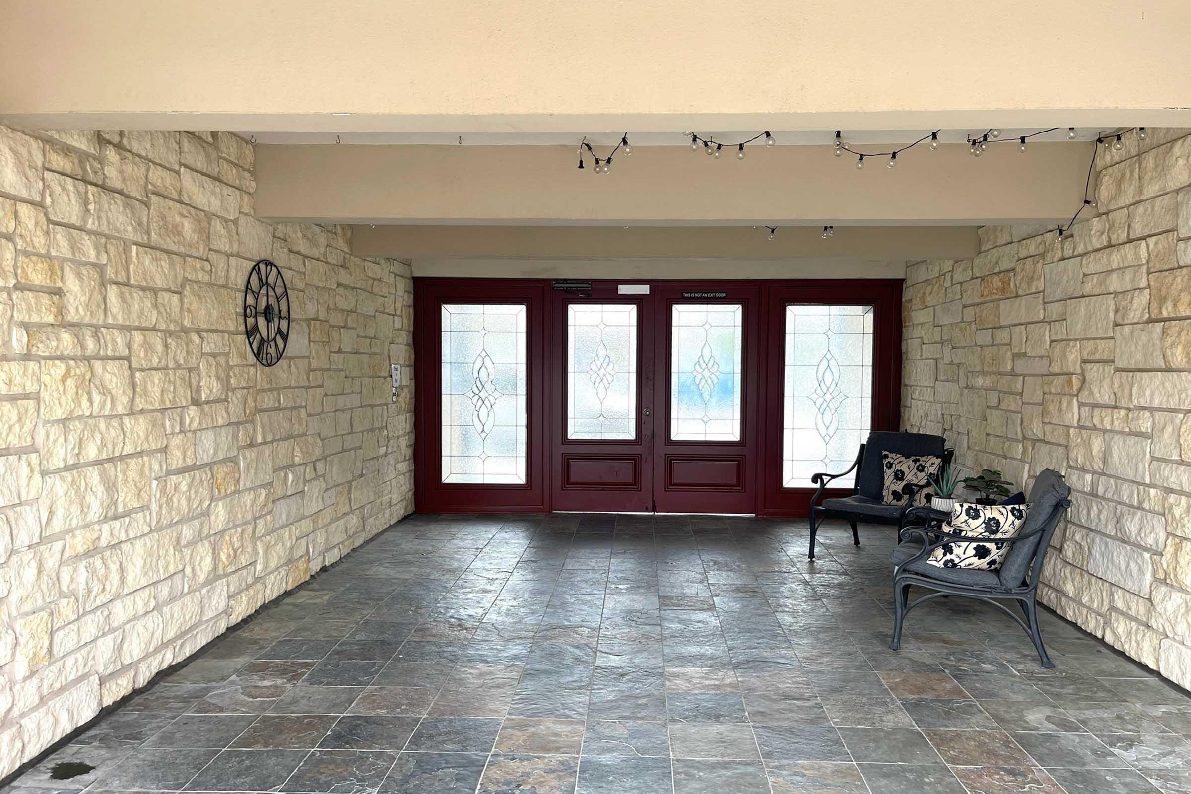 A spacious entryway featuring stone walls, a tiled floor, and double red doors with decorative glass panels. Two chairs with decorative cushions are arranged against the wall, and string lights are hung above, adding a cozy atmosphere.