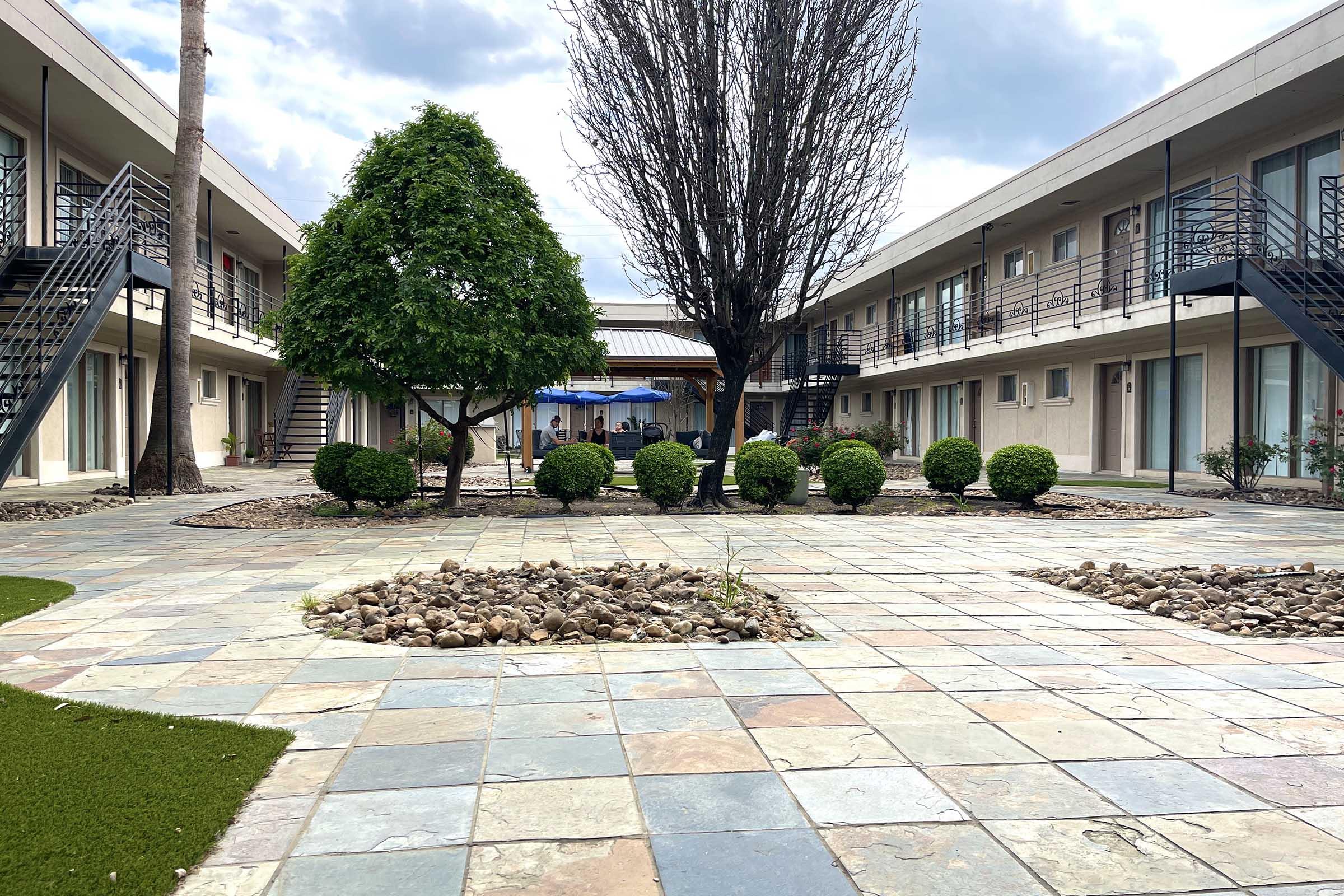A courtyard of a motel featuring a landscaped area with manicured bushes, stone pathways, and decorative rocks. Two stories of exterior corridor rooms are visible, with a tree and an umbrella on the patio in the background under a cloudy sky.
