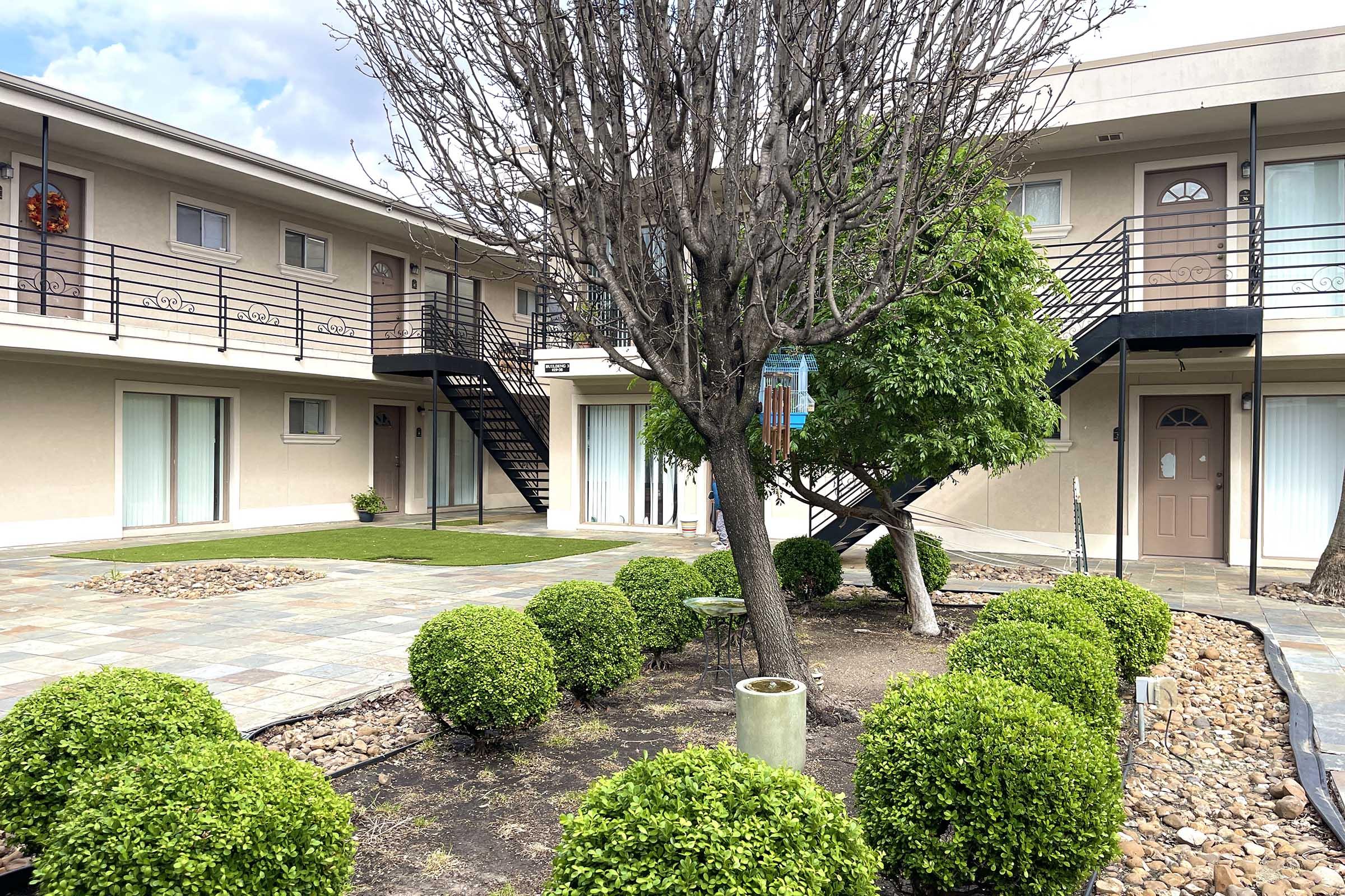 A view of a courtyard at a motel featuring a barren tree, manicured round bushes, and stone pathways. The backdrop includes two levels of the motel with balconies and a staircase, surrounded by green grass and a cloudy sky.