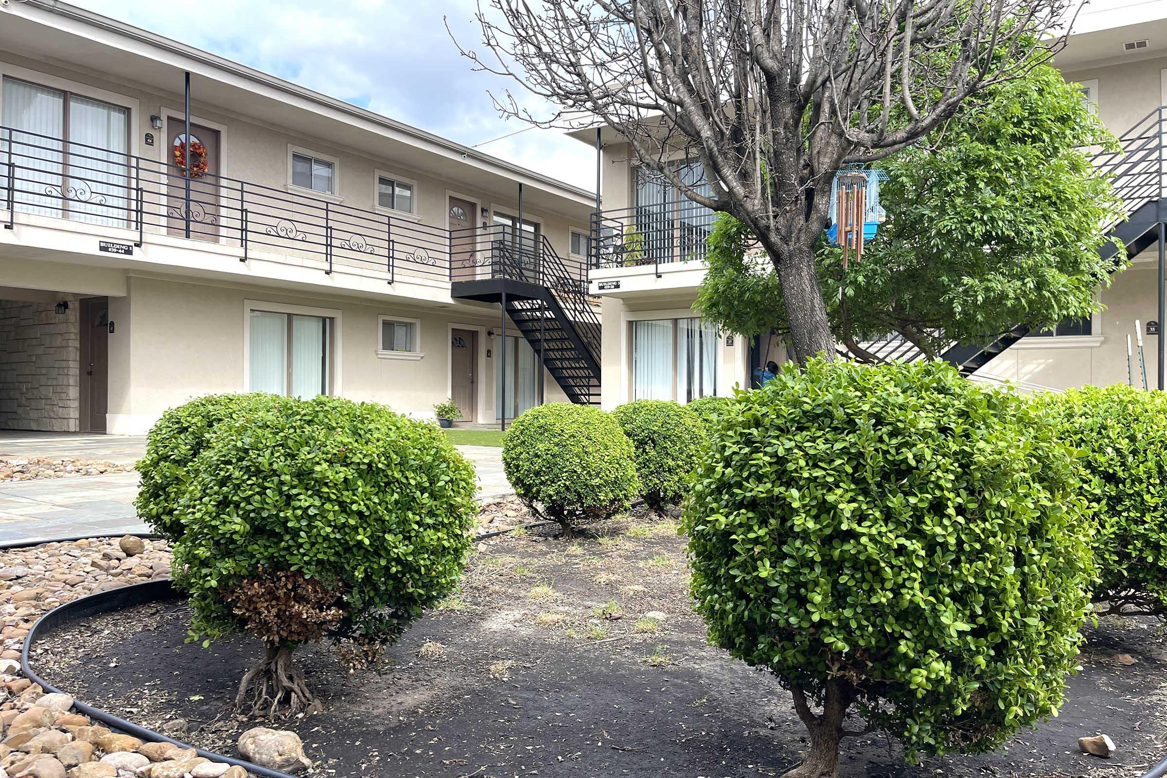 A view of a courtyard in an apartment complex featuring two-story buildings with metal stairs. Neatly trimmed round bushes surround a patch of bare ground, and there are small rocks on the ground. The sky is partly cloudy, and the area appears well-maintained.