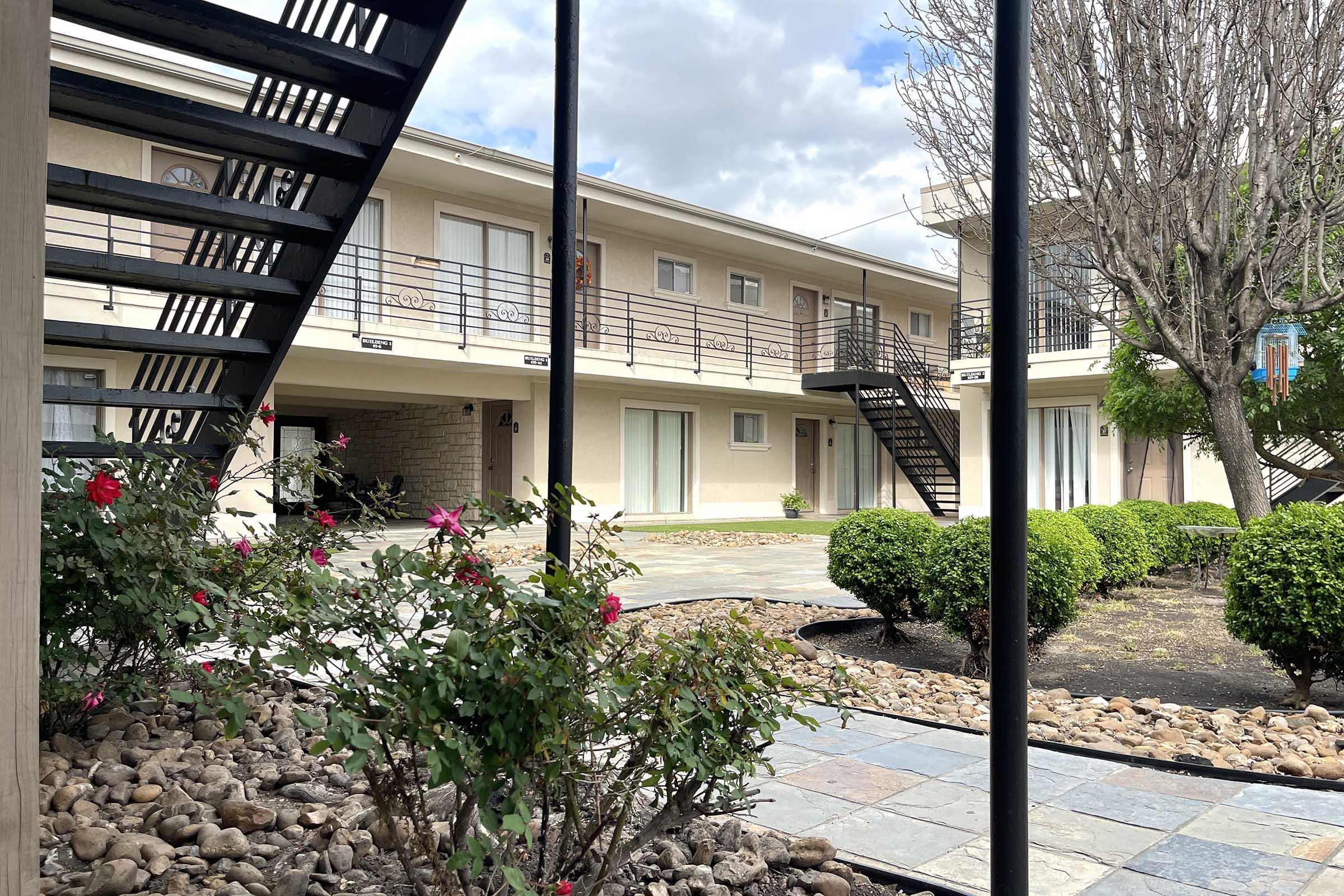 A view of a courtyard in an apartment complex featuring a stone pathway, neatly trimmed shrubs, and blooming rose bushes. Two symmetrical buildings are visible, each with balconies and staircases, under a partly cloudy sky. The scene conveys a peaceful, well-maintained outdoor space.