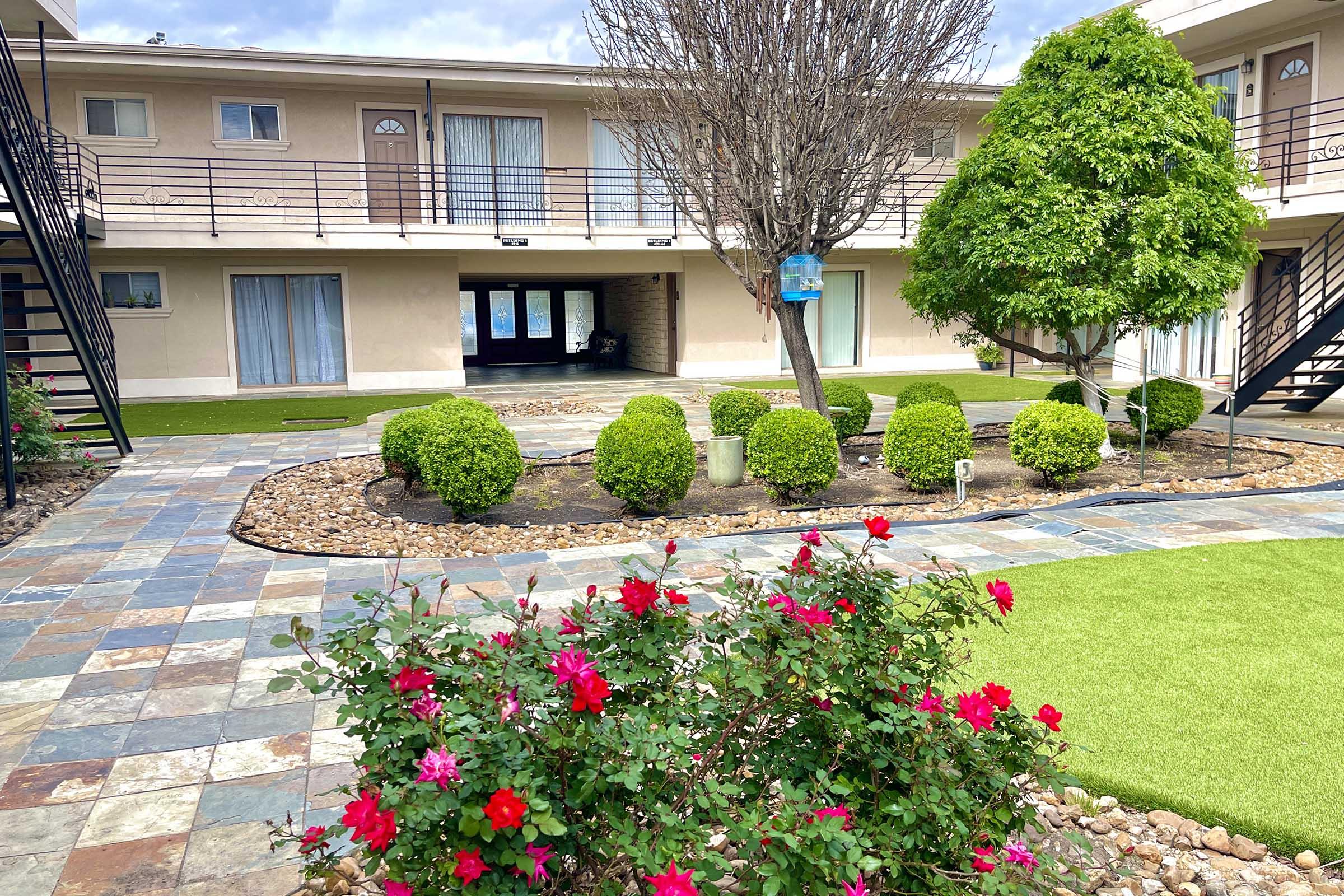 A tranquil courtyard featuring manicured bushes, vibrant red flowers, and a stone path. Two buildings with balconies flank the area, while a tree adds greenery. The scene is bright and inviting, perfect for relaxing outdoors.
