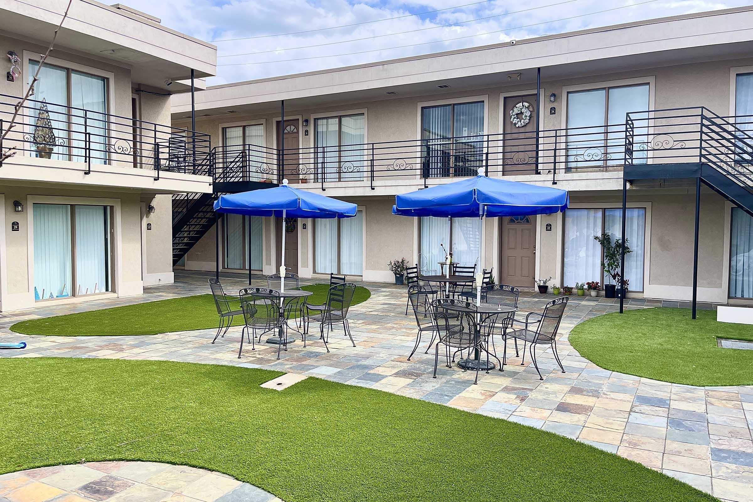 Outdoor courtyard of a motel featuring round tables and chairs under blue umbrellas. The area is decorated with artificial grass and tiled pathways. Stairs lead to upper-level rooms, which have large windows and small plants on the ledges. The sky is partly cloudy, creating a bright and inviting atmosphere.