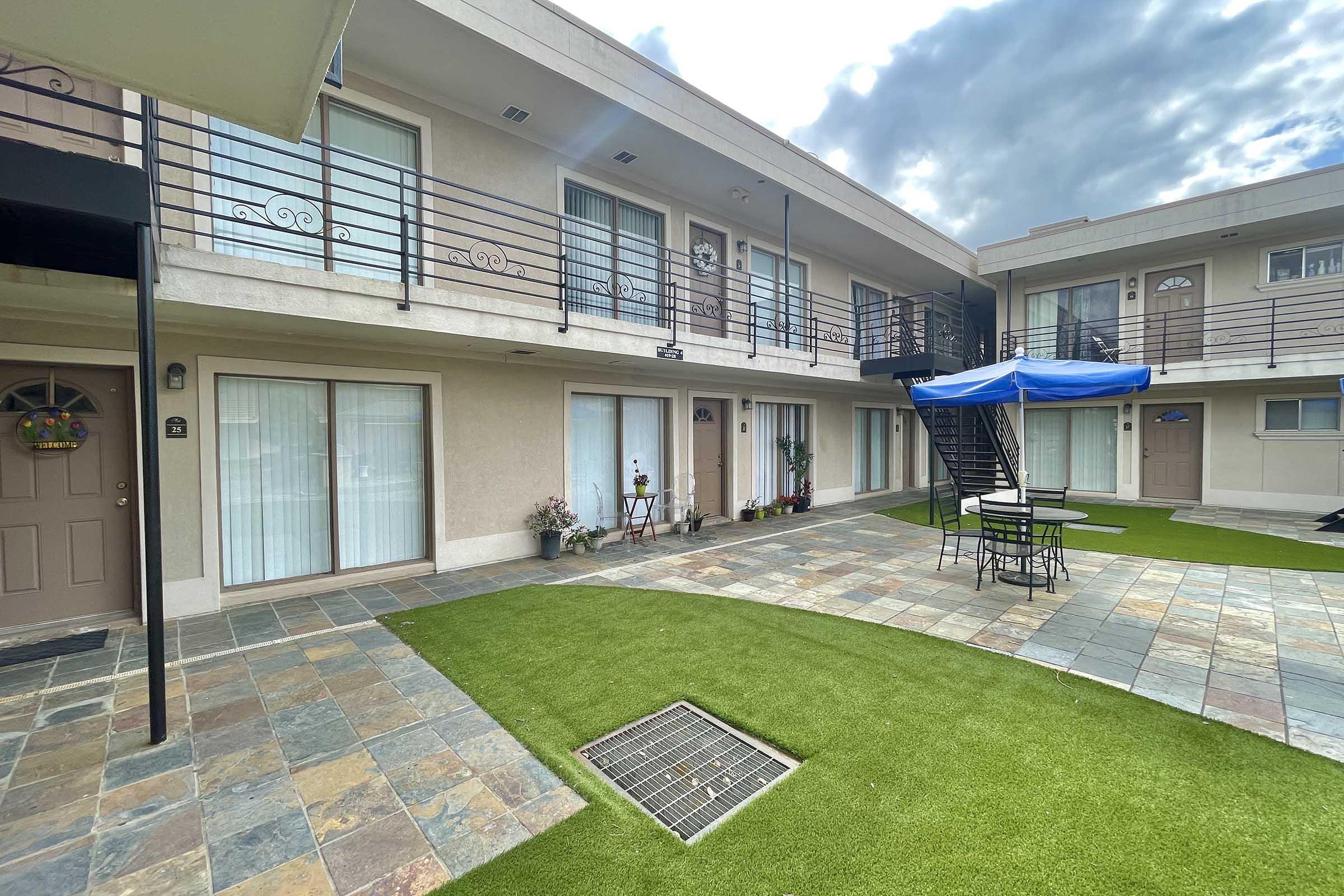 A view of a small courtyard area at a motel, featuring several rooms with doors leading outside. There is a blue umbrella over a table and chairs, surrounded by artificial grass and decorative plants. A staircase leads to the upper floor, and the sky is partly cloudy.