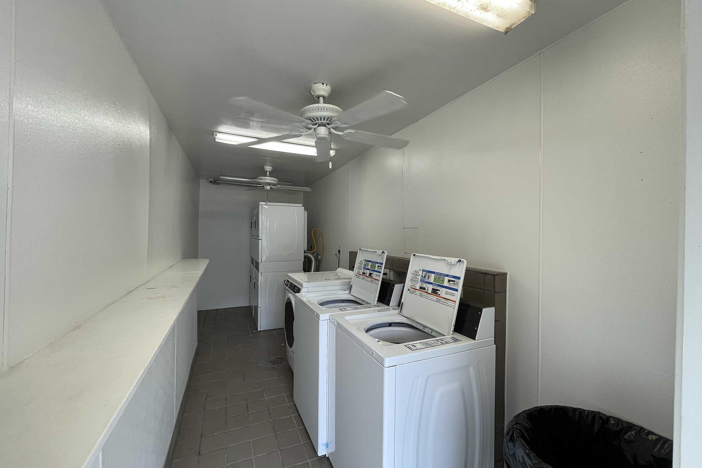 A clean and bright laundry room featuring two washing machines and a dryer. There is a ceiling fan for ventilation, a countertop along one wall, a trash bin in the corner, and a refrigerator in the background. The walls are plain and white, providing a simple, functional space.