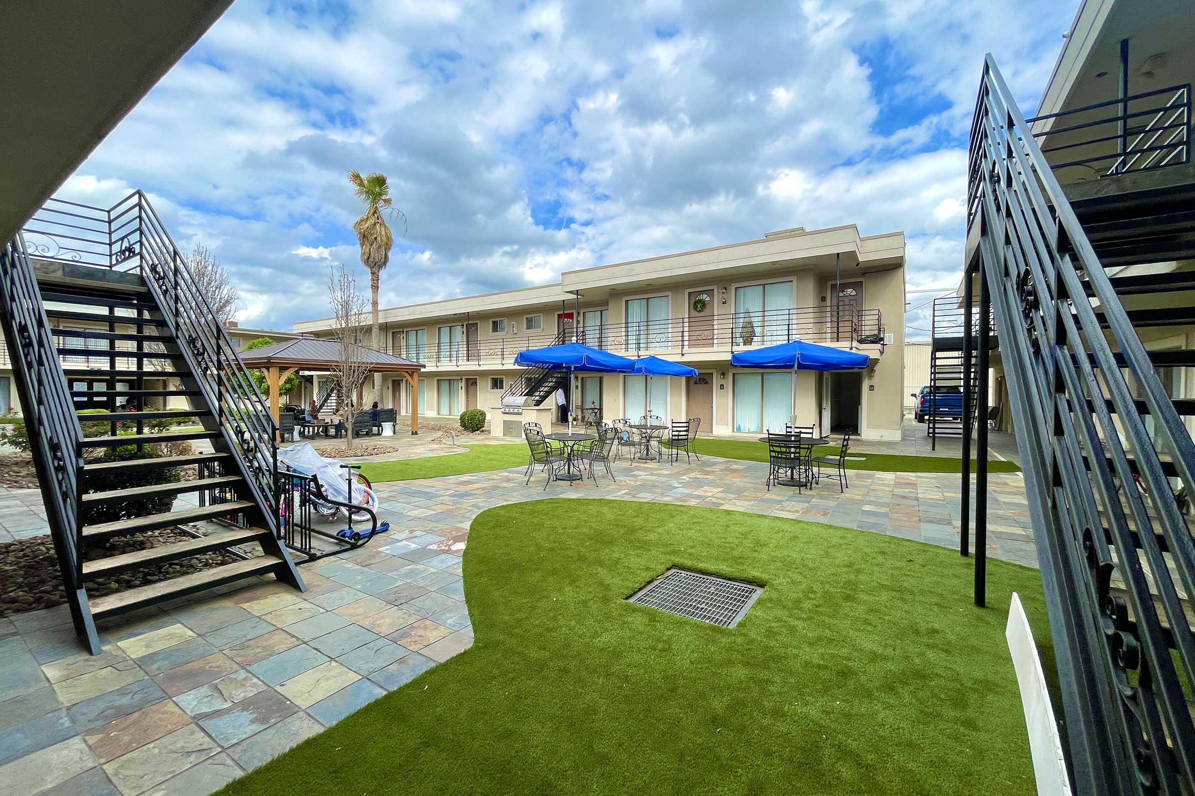 A courtyard of a motel featuring green grass, tables with umbrellas, and a staircase leading to upper floors. The buildings have a modern design and are surrounded by palm trees under a partly cloudy sky.