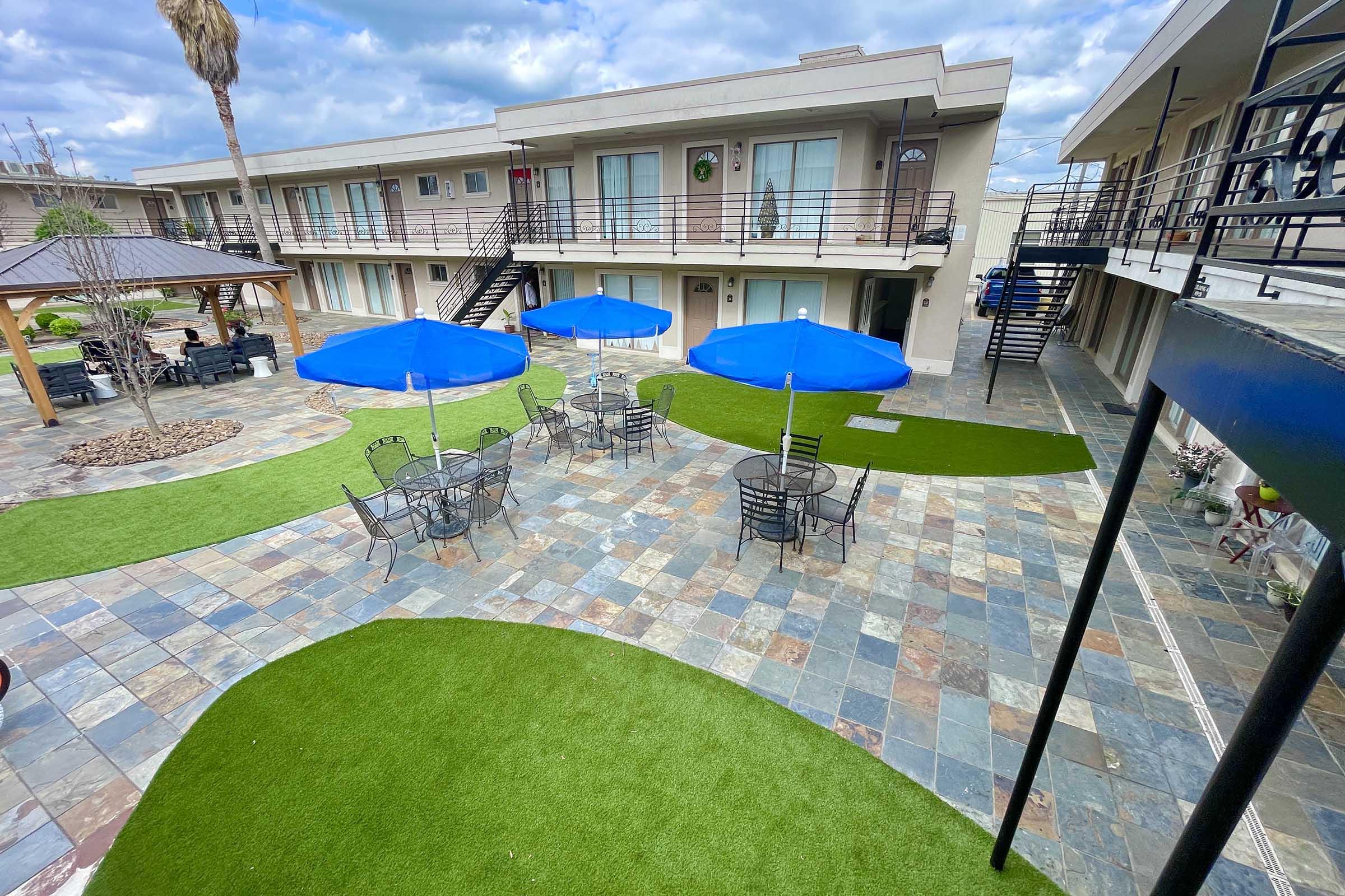A courtyard of a motel featuring a tiled patio with artificial grass, blue umbrella-shaded tables, and chairs. There are palm trees and a gazebo in the background, along with motel rooms that have balconies. The sky is partly cloudy, creating a bright but overcast atmosphere.