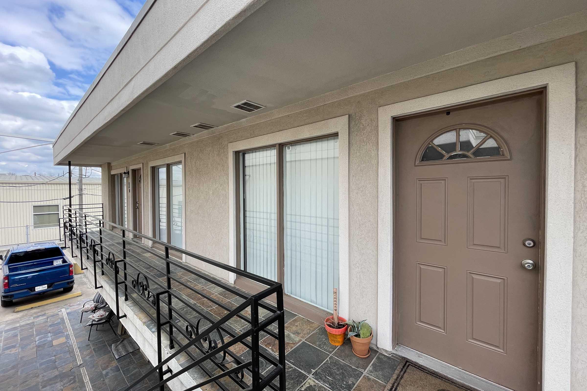 Exterior view of a multi-unit building featuring a light-colored facade, a brown door, and small potted plants near the entrance. A balcony with black railings is visible, along with a portion of a blue vehicle parked nearby. The sky is partly cloudy.