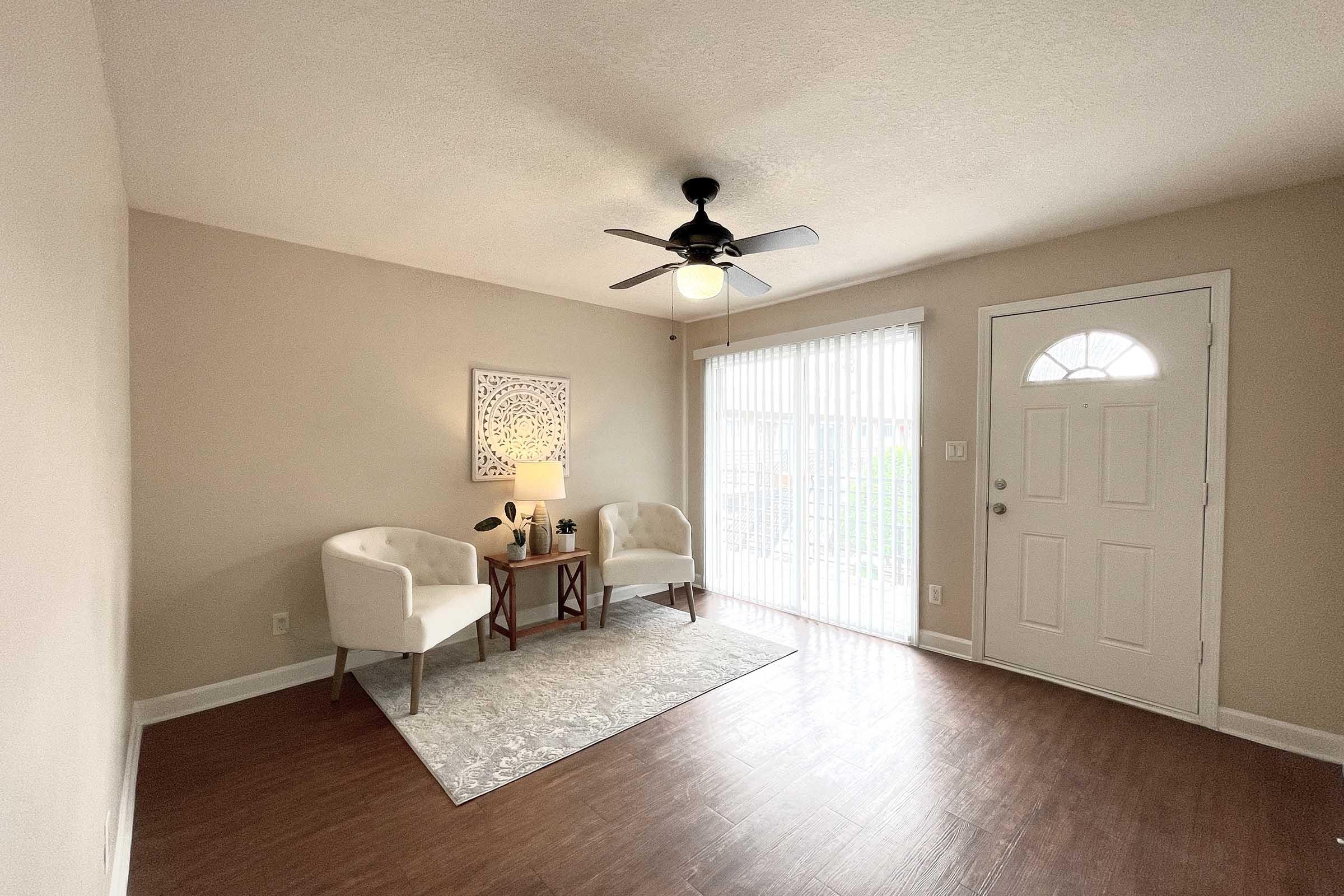 A well-lit living room featuring two white armchairs facing each other on a light rug, a small wooden table with a lamp in between, and a decorative wall piece. There's a door with a window and blinds that leads to an outdoor area, and stylish wooden flooring enhances the cozy atmosphere.