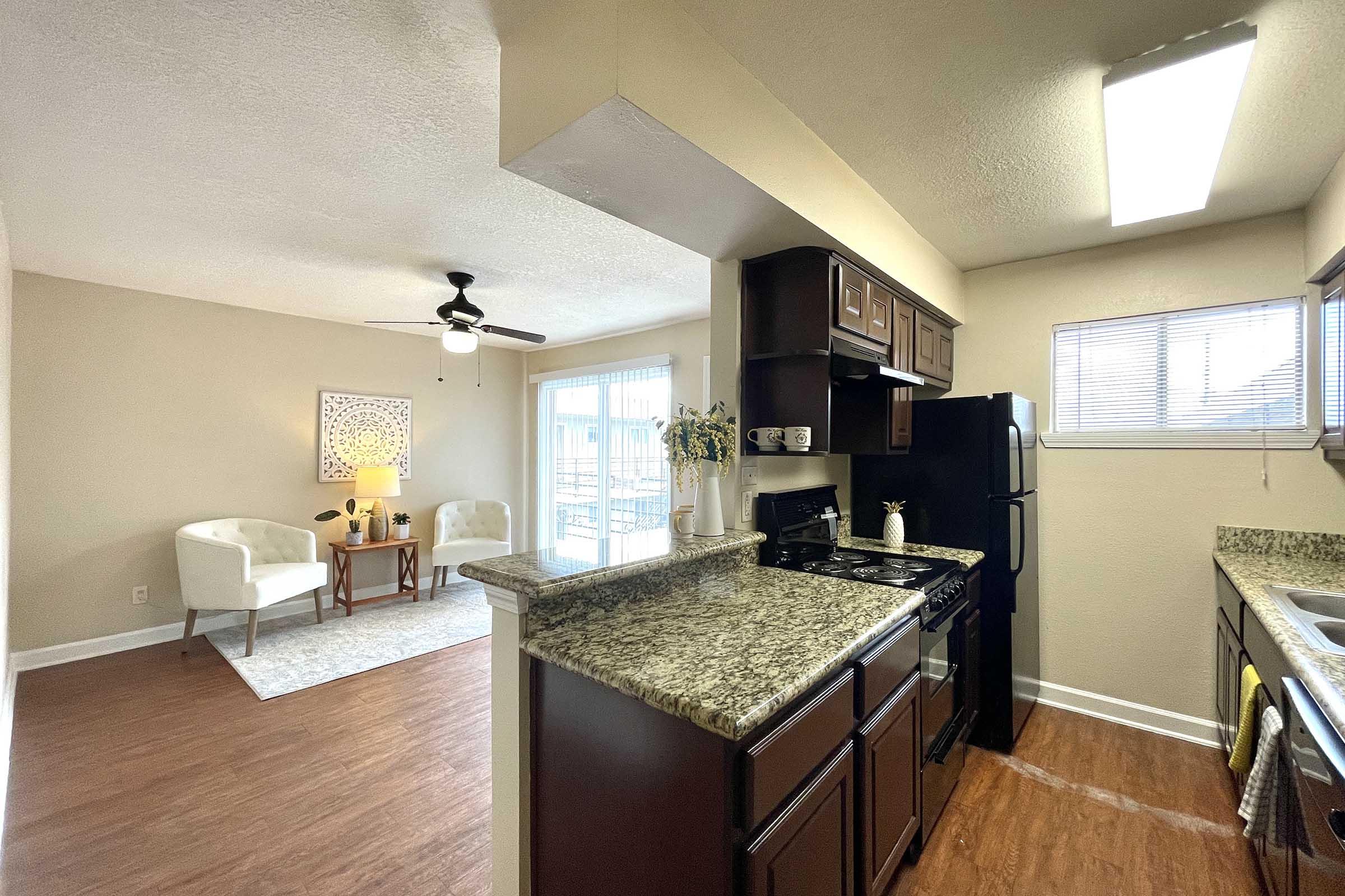 A modern kitchen with granite countertops, dark wooden cabinets, and black appliances, including a stove and refrigerator. Adjacent is a cozy living area with two white chairs and a decorative rug, illuminated by natural light from a window. The space has neutral wall colors and a comforting ambiance.