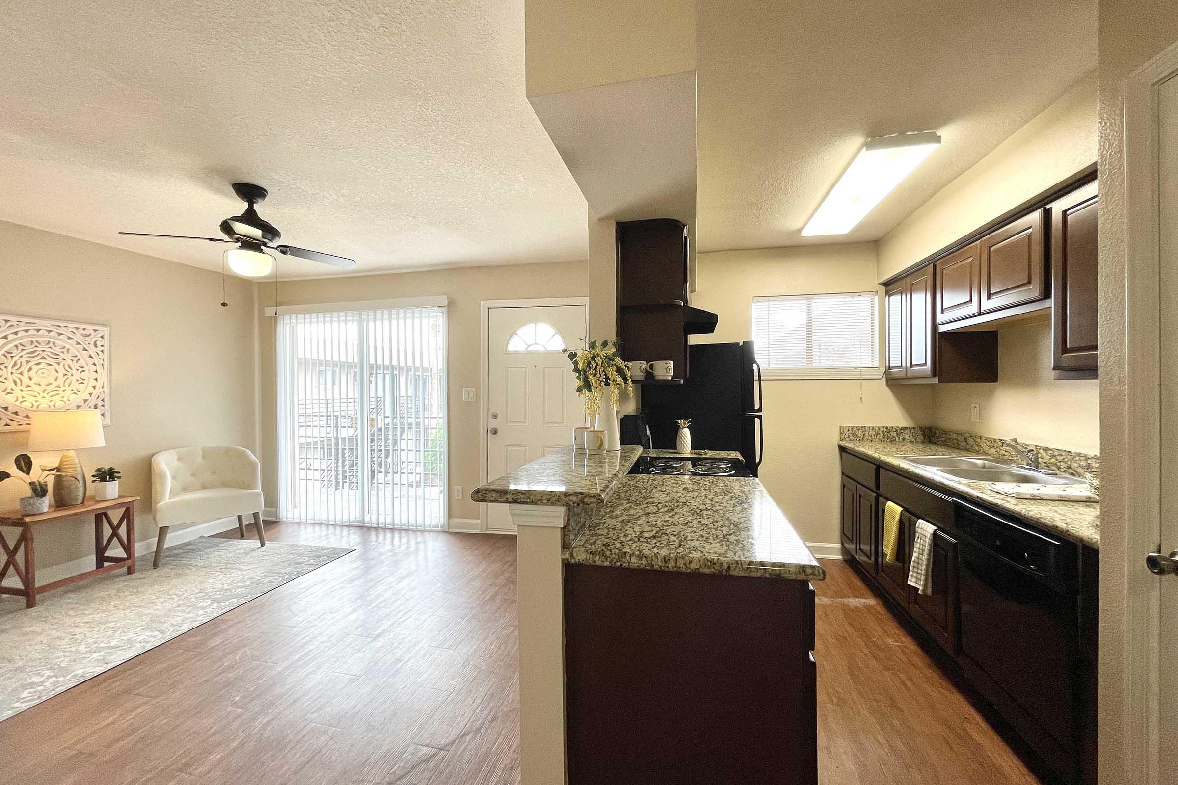 Open concept living space featuring a modern kitchen with dark cabinets and granite countertops, a dining area with a ceiling fan, and a cozy living area with a sofa. Natural light streams in from sliding glass doors leading to an outdoor patio.