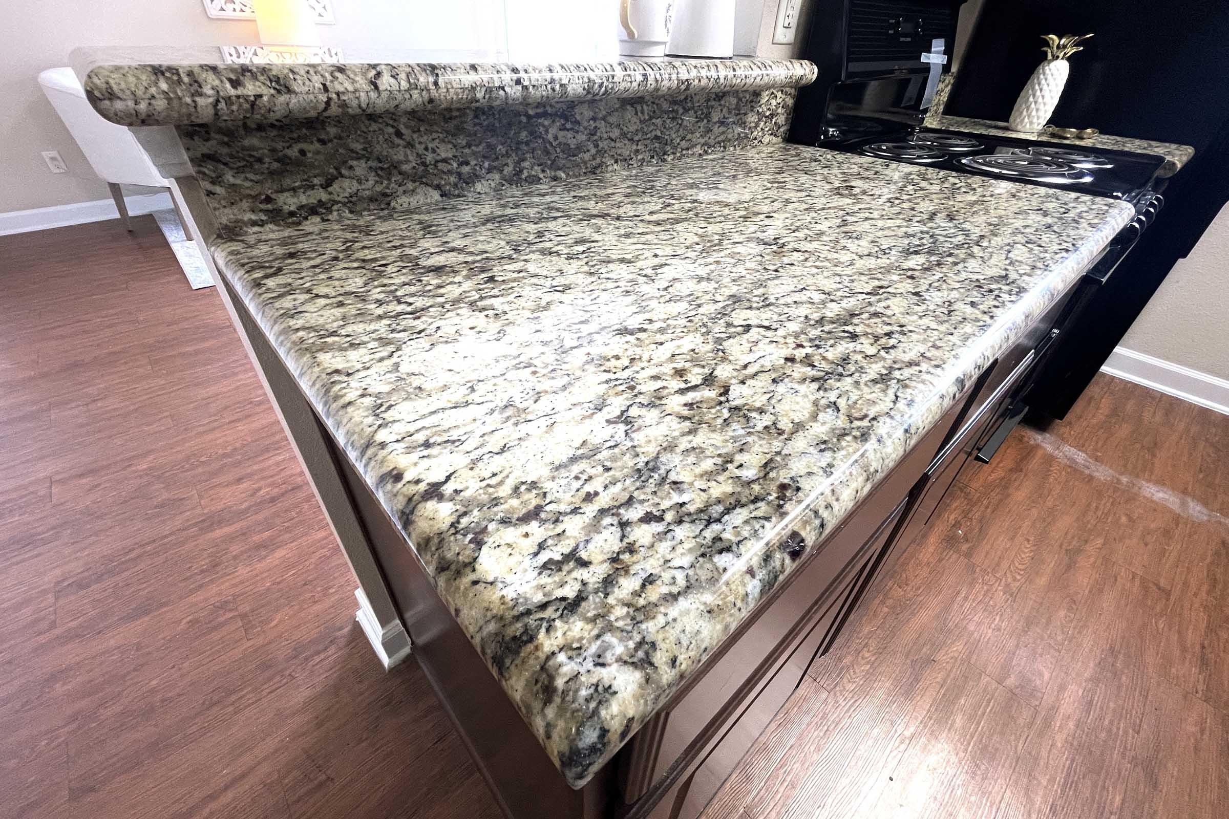 Close-up view of a kitchen countertop featuring a granite surface with a mix of beige, brown, and grey tones. The countertop has a polished finish and edges, and there is a portion of a kitchen stove visible in the background, along with light-colored cabinetry and an area to the left.