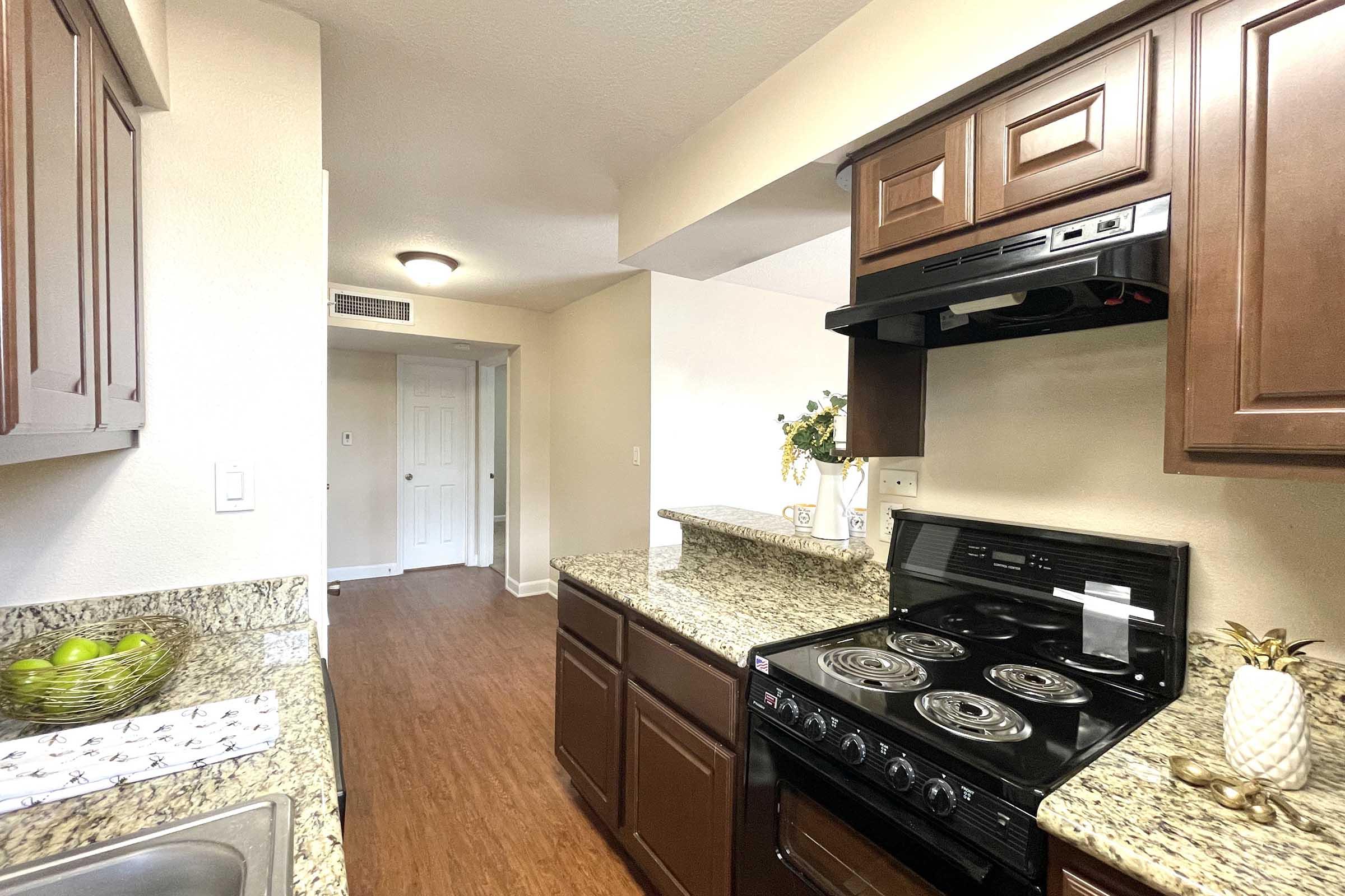 A kitchen featuring dark wood cabinetry, granite countertops, and a black stove with an overhead microwave. The sink area includes a bowl of green apples, and there is a doorway leading to another room in the background, illuminated by natural light. The floors are wooden, creating a warm atmosphere.