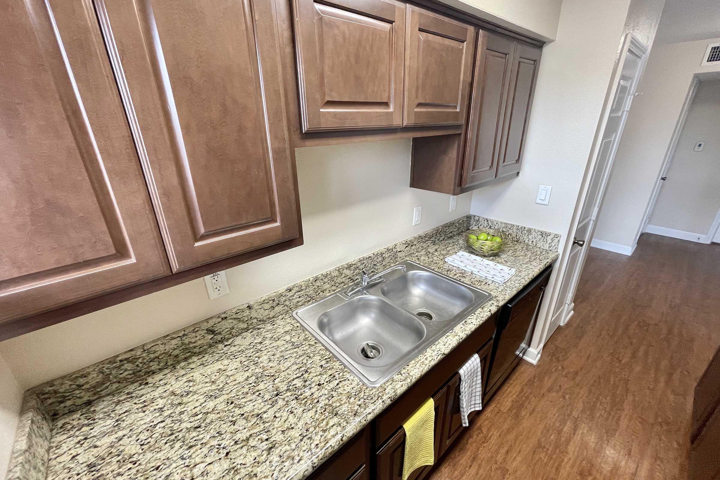 A modern kitchen featuring brown wooden cabinets above a granite countertop. Two stainless steel sinks are visible, along with a small dish drying rack. A bowl of green fruit sits on the counter, and light-colored towels are hanging from a rack. The flooring is wood-like laminate, and a doorway leads to another room in the background.