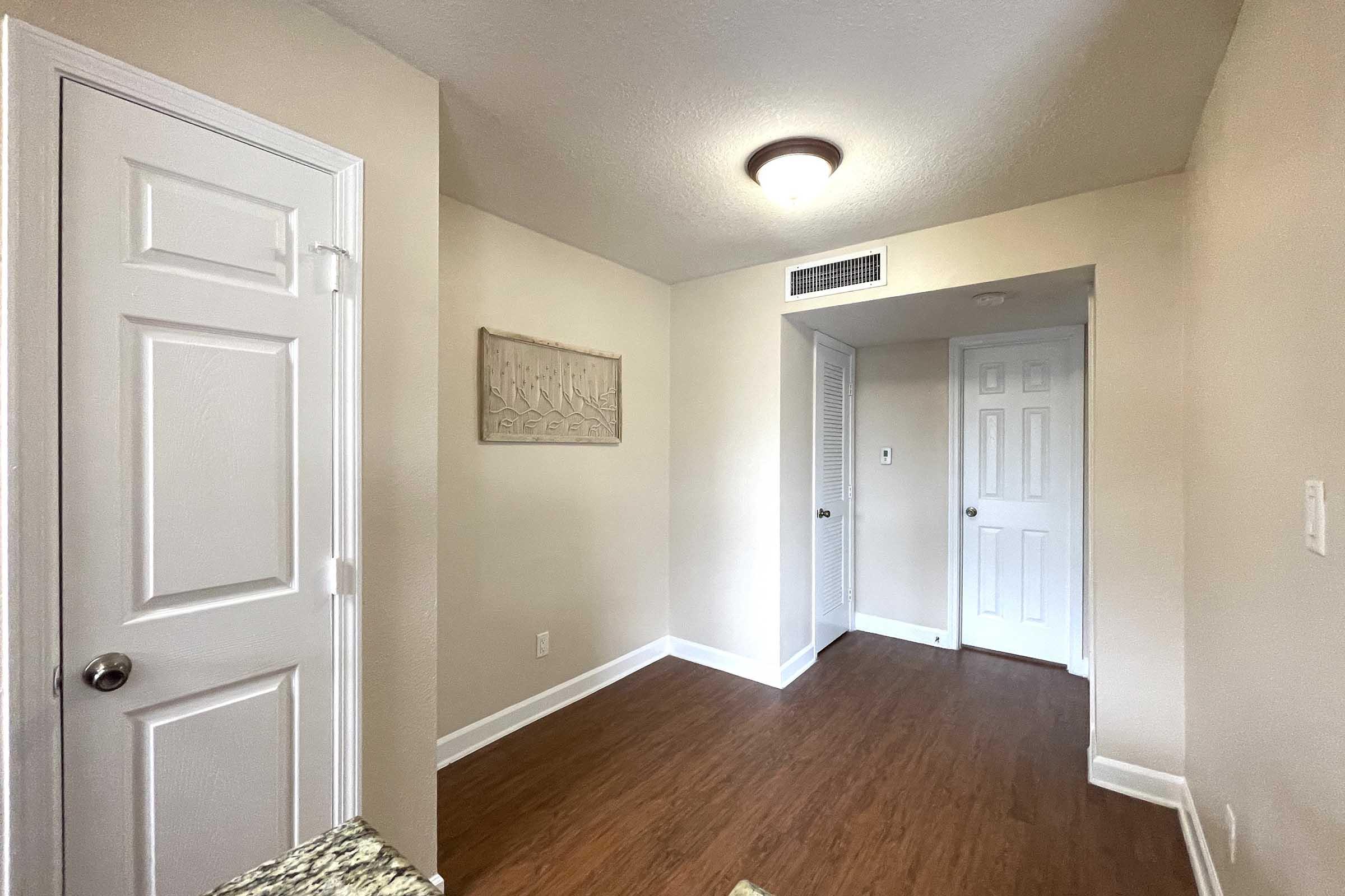 A well-lit interior space featuring light-colored walls and wood flooring. On the left, there is a closed white door, and to the right, a doorway leads to another room with two closed doors. A decorative wall panel is mounted on the wall. The overall atmosphere is clean and inviting.