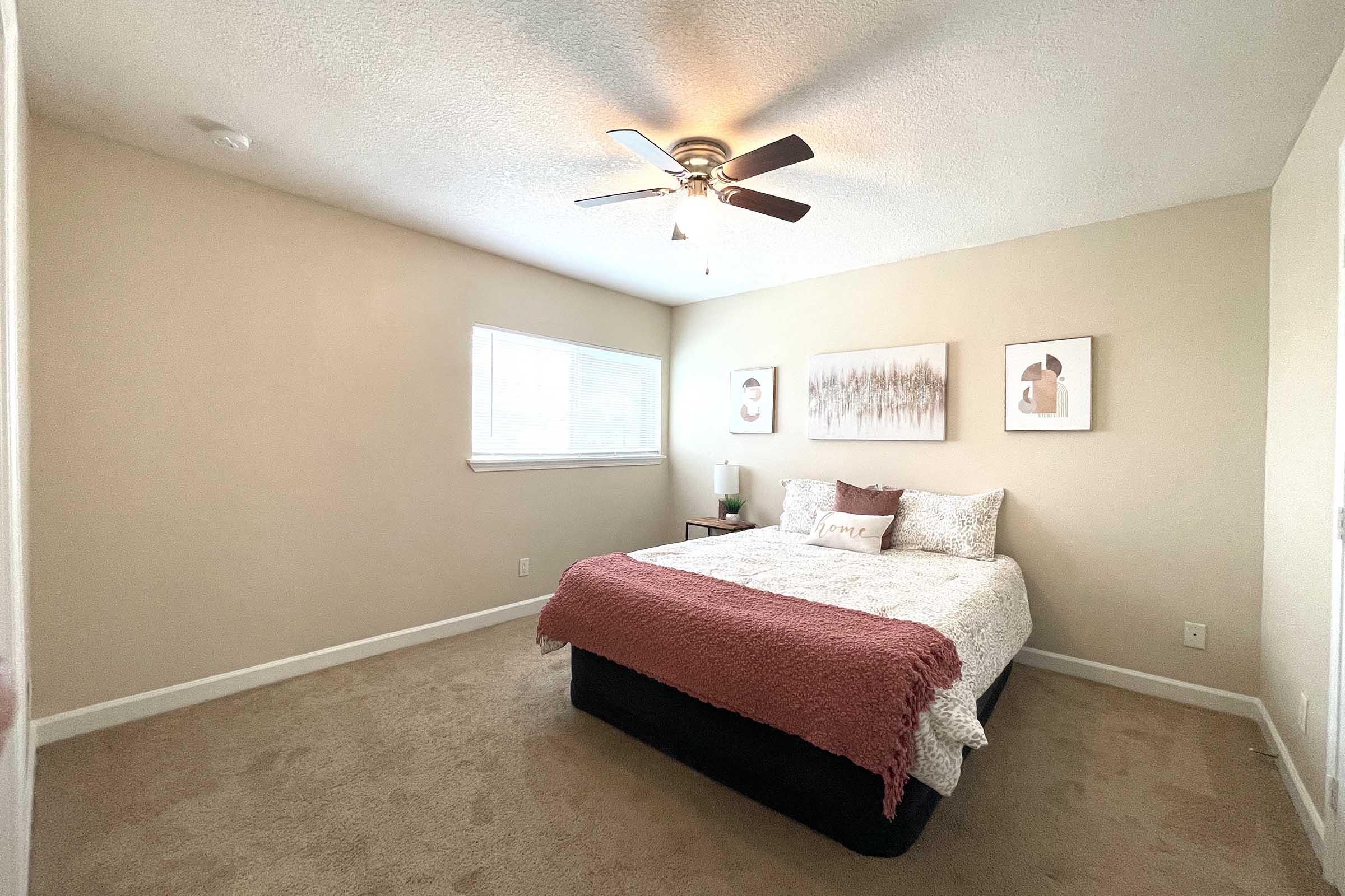 A cozy bedroom featuring a bed with a light-colored quilt and a maroon throw blanket. There are two framed art pieces on the wall, a nightstand with a lamp beside the bed, and a ceiling fan overhead. The room is well-lit with natural light coming through a window, and the walls are painted in a soft, neutral tone.