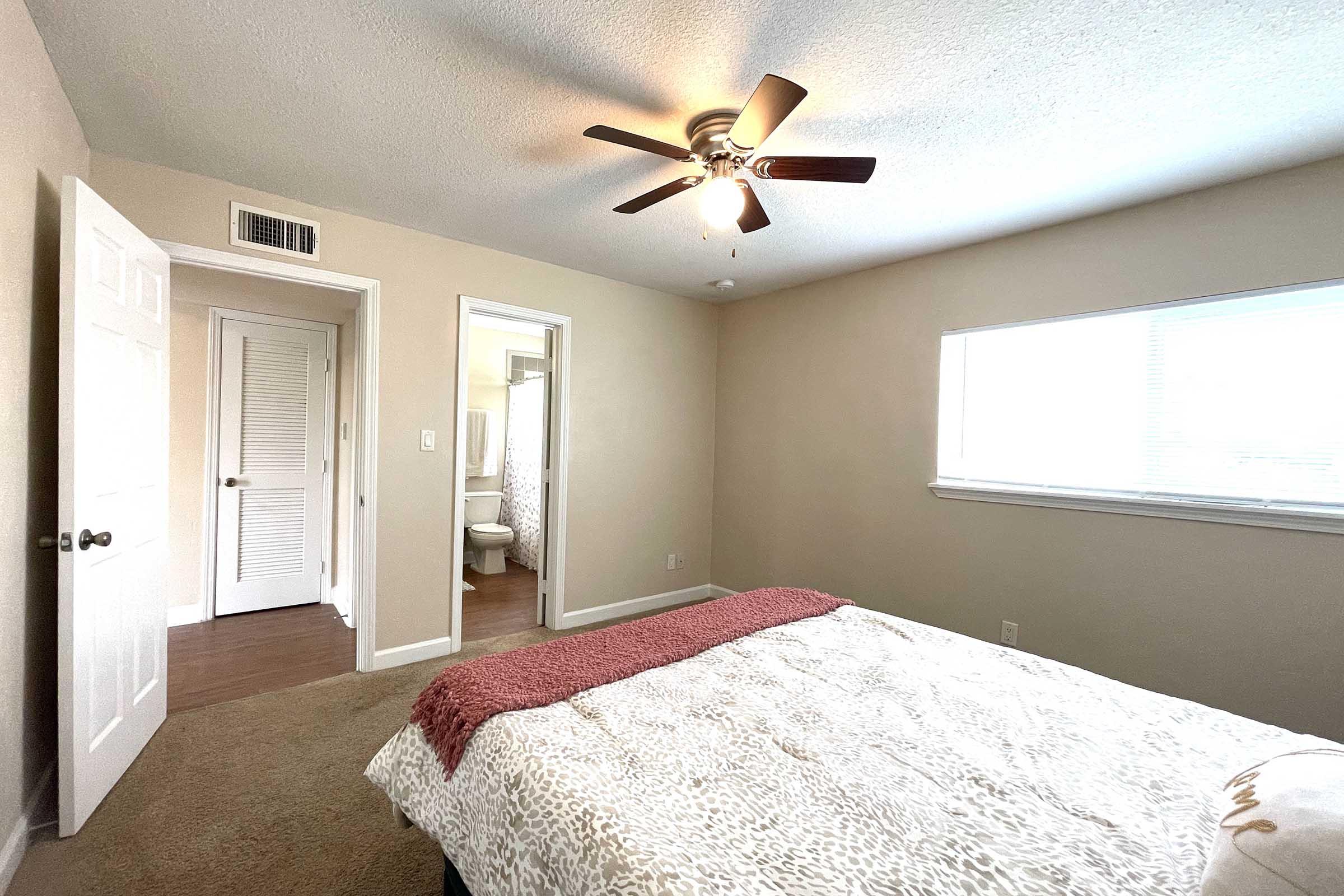 A cozy bedroom featuring a light-colored wall, a ceiling fan with wooden blades, and a neatly made bed with a patterned blanket. There are two doors leading to other rooms, one likely a bathroom, and a window allowing natural light to brighten the space. The flooring is a warm, wood-like material.