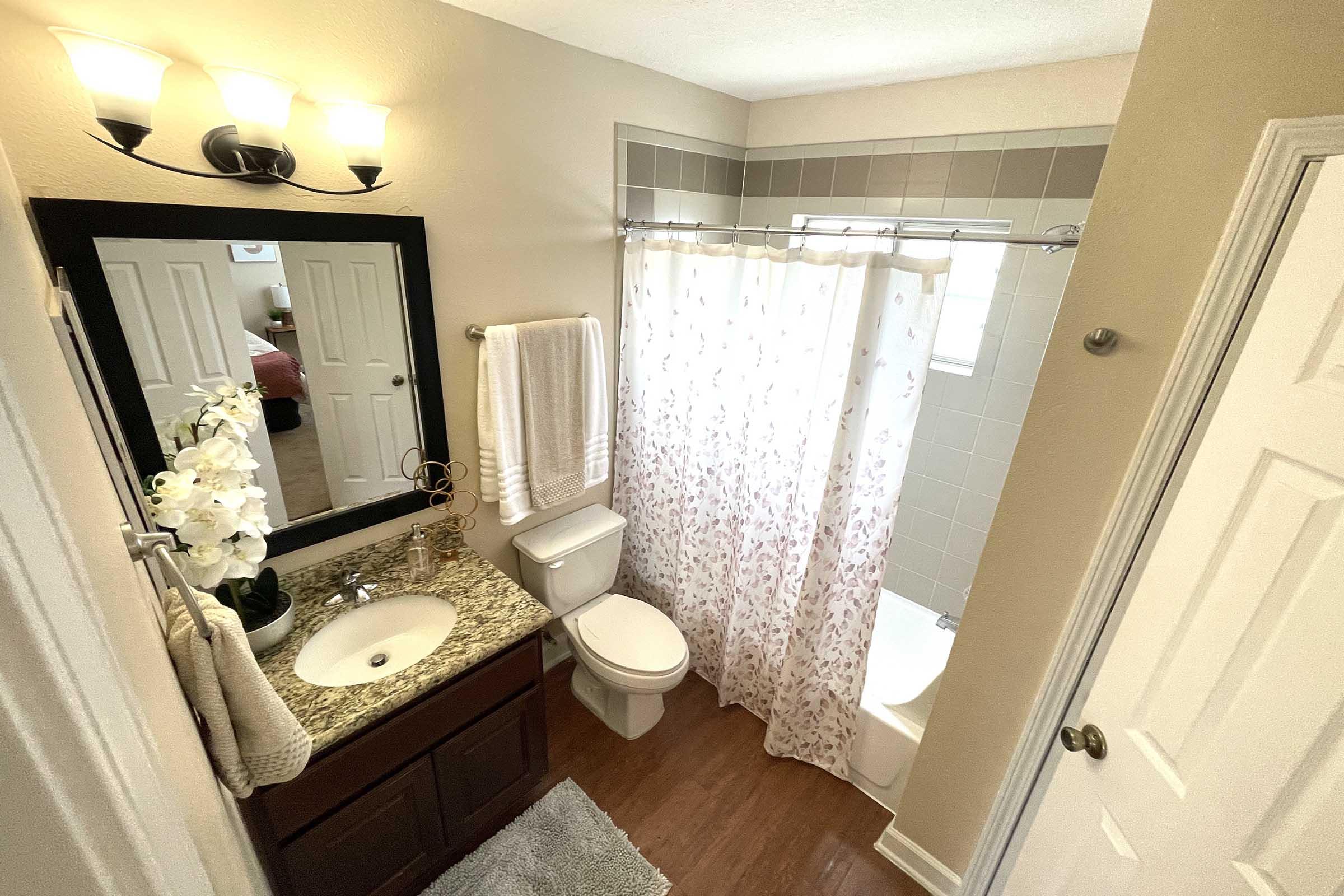 A well-lit bathroom featuring a granite countertop with a sink, a large mirror, and a decorative orchid. The toilet and a shower with a floral curtain are also visible, along with a soft rug on the floor. Light-colored walls contribute to a clean and inviting atmosphere.
