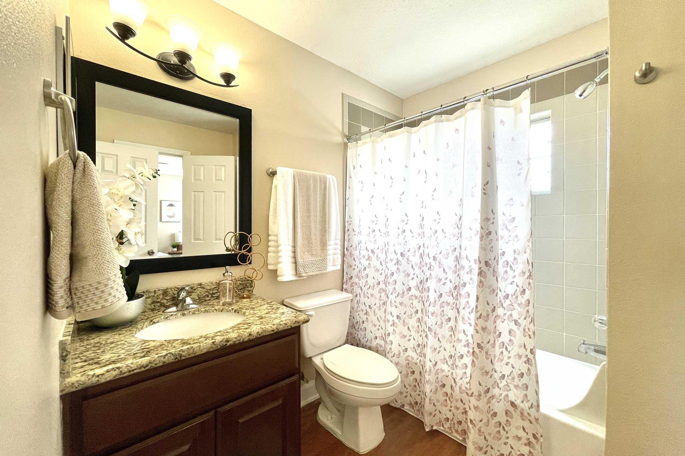 A well-lit bathroom featuring a granite countertop with a sink, a large mirror framed in black, a white shower curtain with floral patterns, a toilet, and towels hanging on a rack. The space is clean and inviting, with a neutral color scheme and natural lighting.