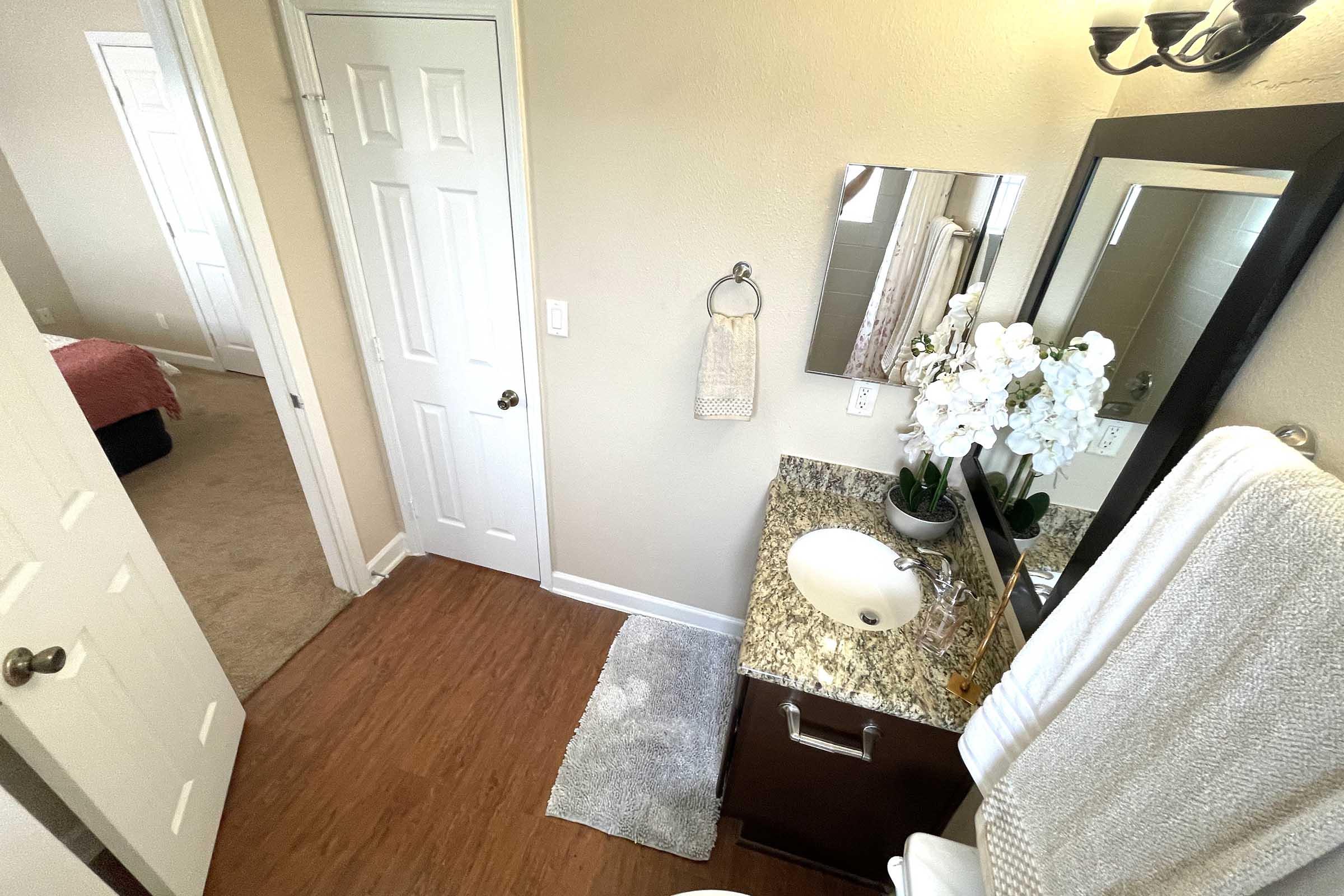 A well-lit bathroom featuring a granite countertop sink, a mirror mounted on the wall, and a decorative arrangement of white flowers. There is a towel rack and a plush towel hanging nearby. The room has a light-colored wall and an adjoining room partially visible, with a carpeted area.