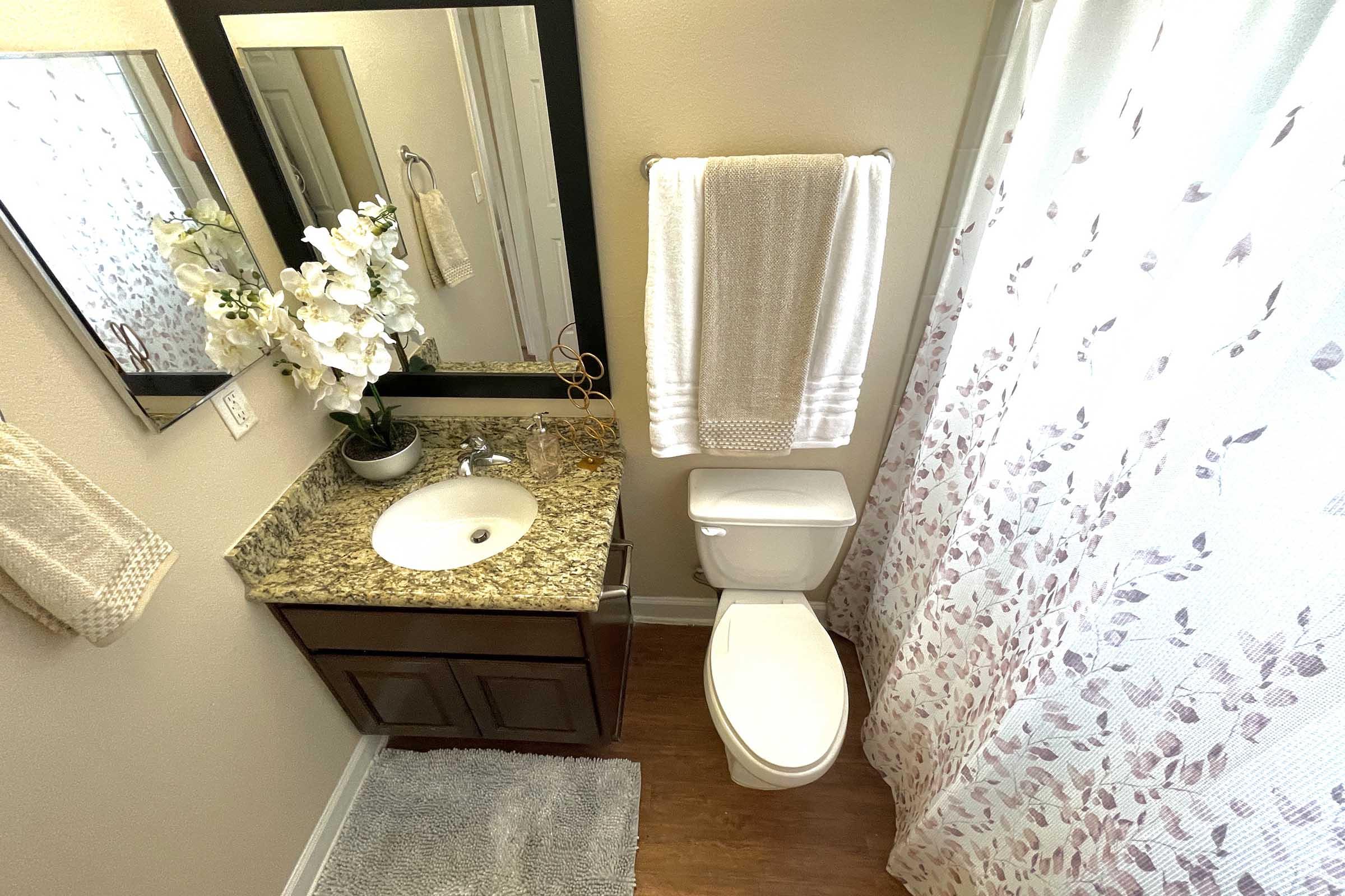 A well-lit bathroom featuring a granite countertop with a sink, a mirror above, and decorative items like a plant and flowers. A white towel hangs nearby, and a shower curtain with a floral design is visible. The toilet is positioned next to the sink, with a plush rug on the floor.